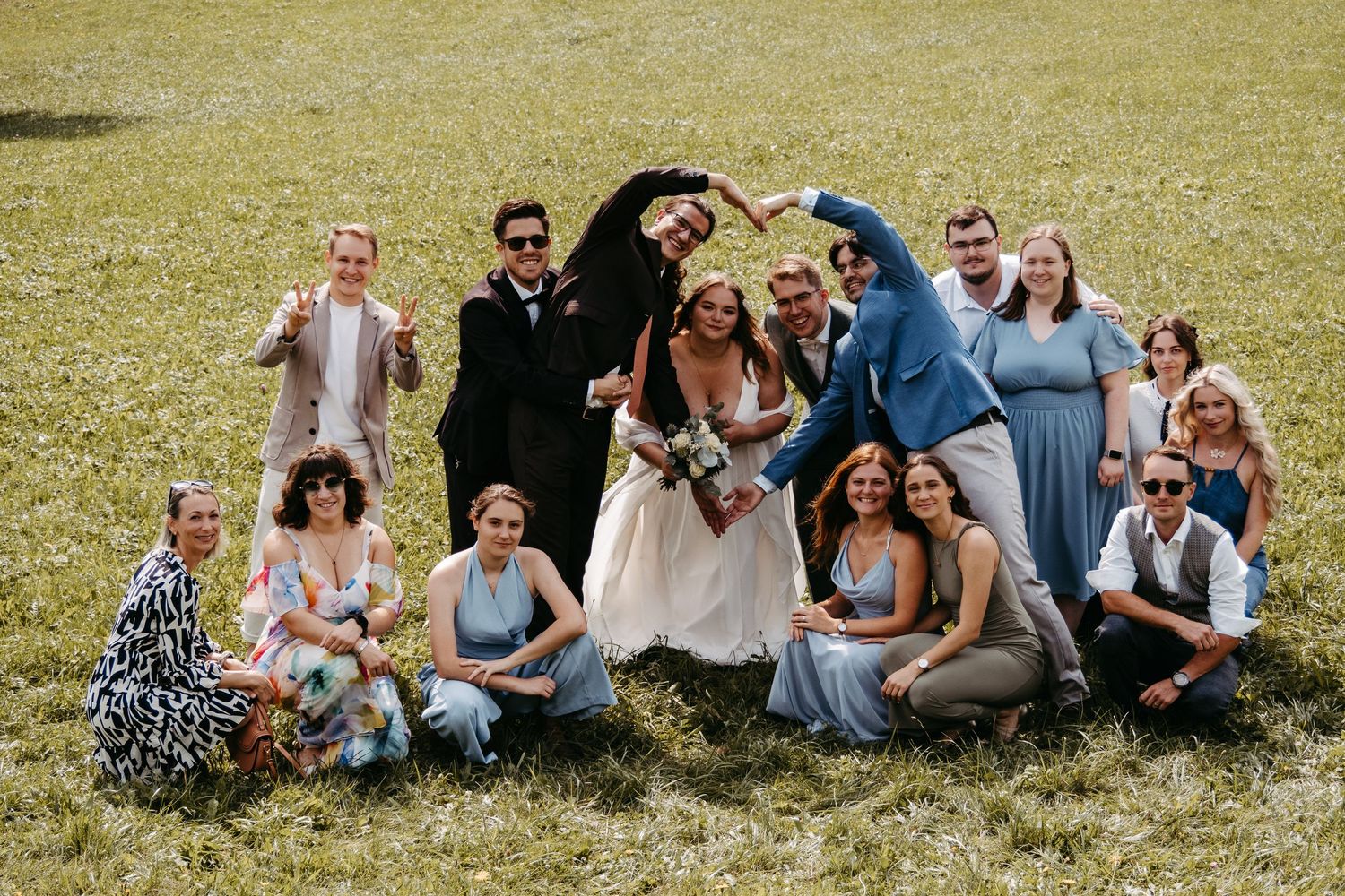 Wedding party celebrating outdoors on green grass, bride and groom surrounded by bridesmaids and groomsmen posing joyfully.