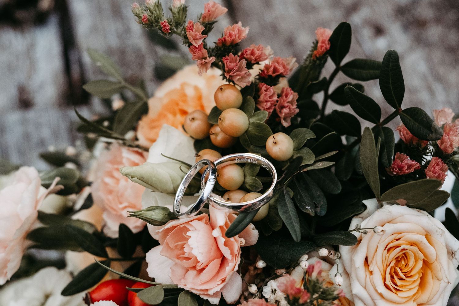 Two silver wedding rings rest on a romantic bridal bouquet of peach roses, pink flowers, and greenery.