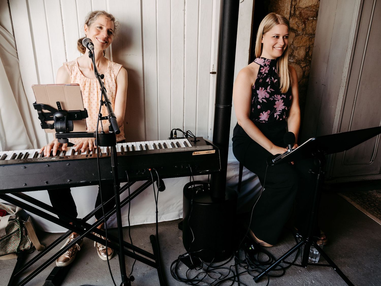 Two female musicians performing, one playing keyboard and one with flute, in a rustic indoor venue.