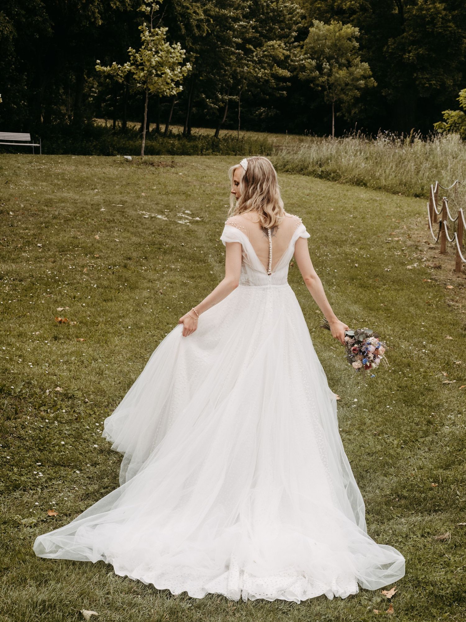 Bride in elegant off-shoulder white ball gown with long train holds bouquet on lush green lawn.
