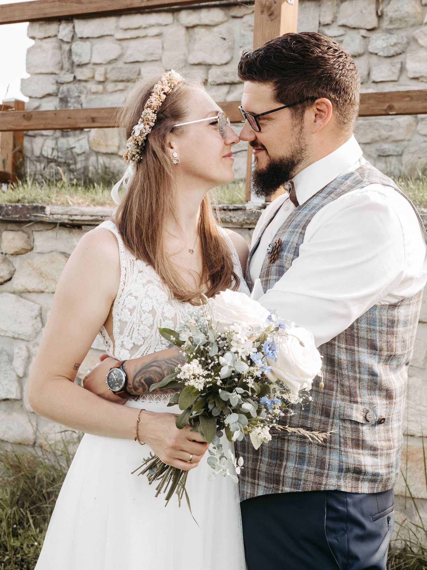 A bride and groom share a romantic moment, she holds a wildflower bouquet wearing a floral crown, he wears a plaid vest.