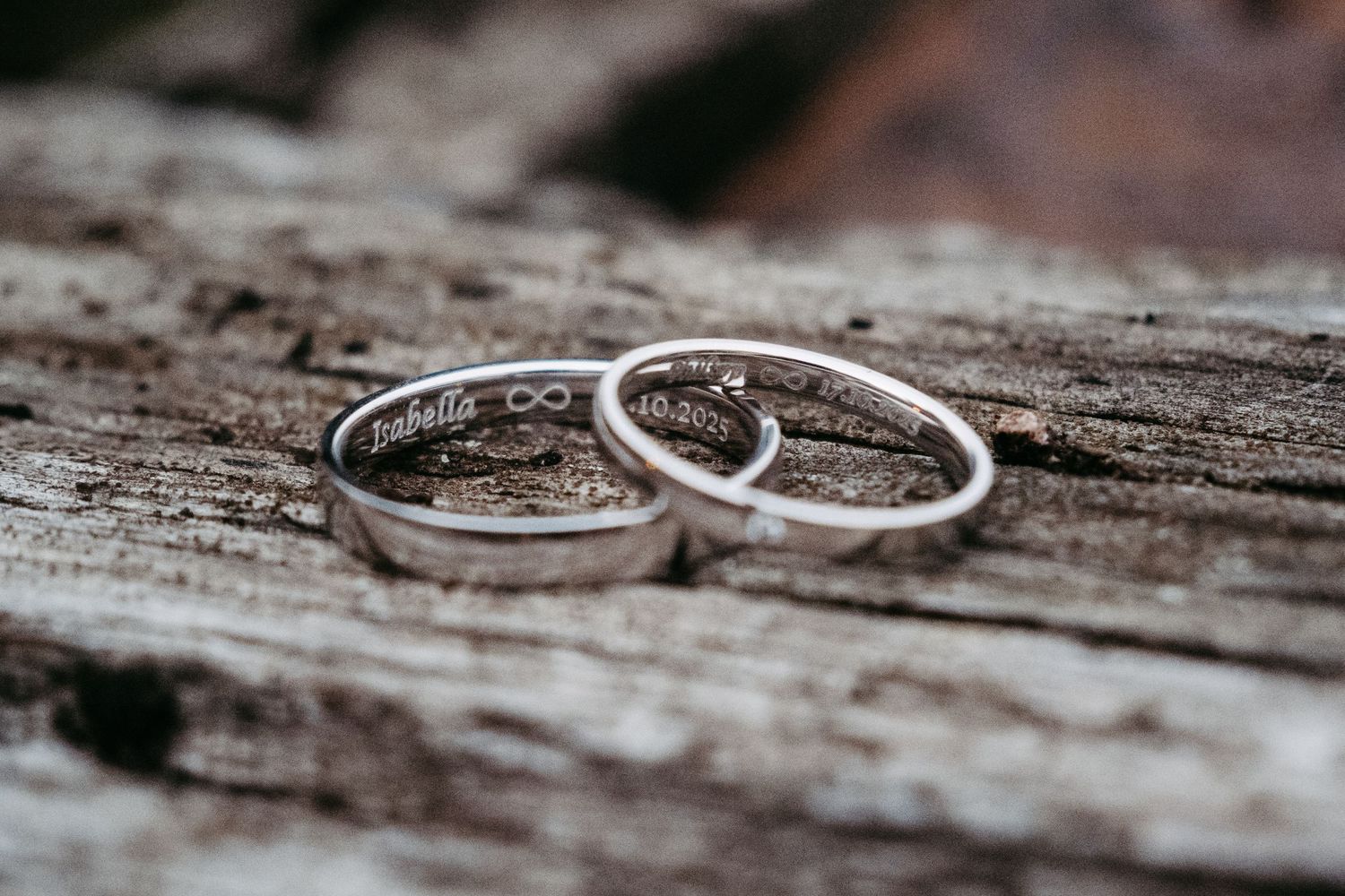 Two engraved silver wedding bands resting on rustic wood, inscribed with names and a date.