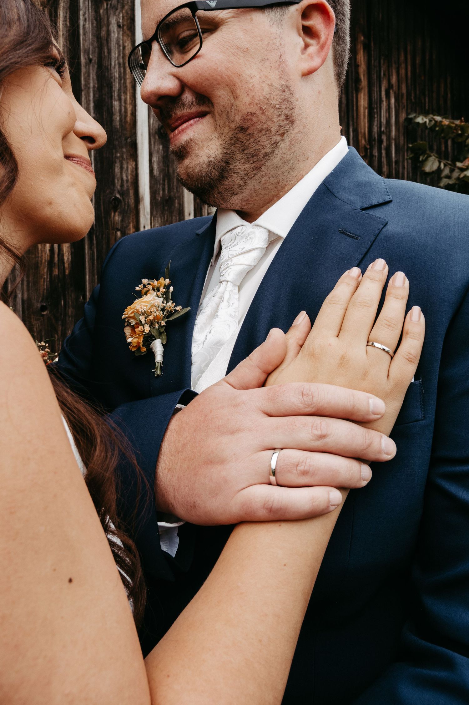 Close-up of bride and groom showing wedding rings, groom wearing navy suit with floral boutonniere.