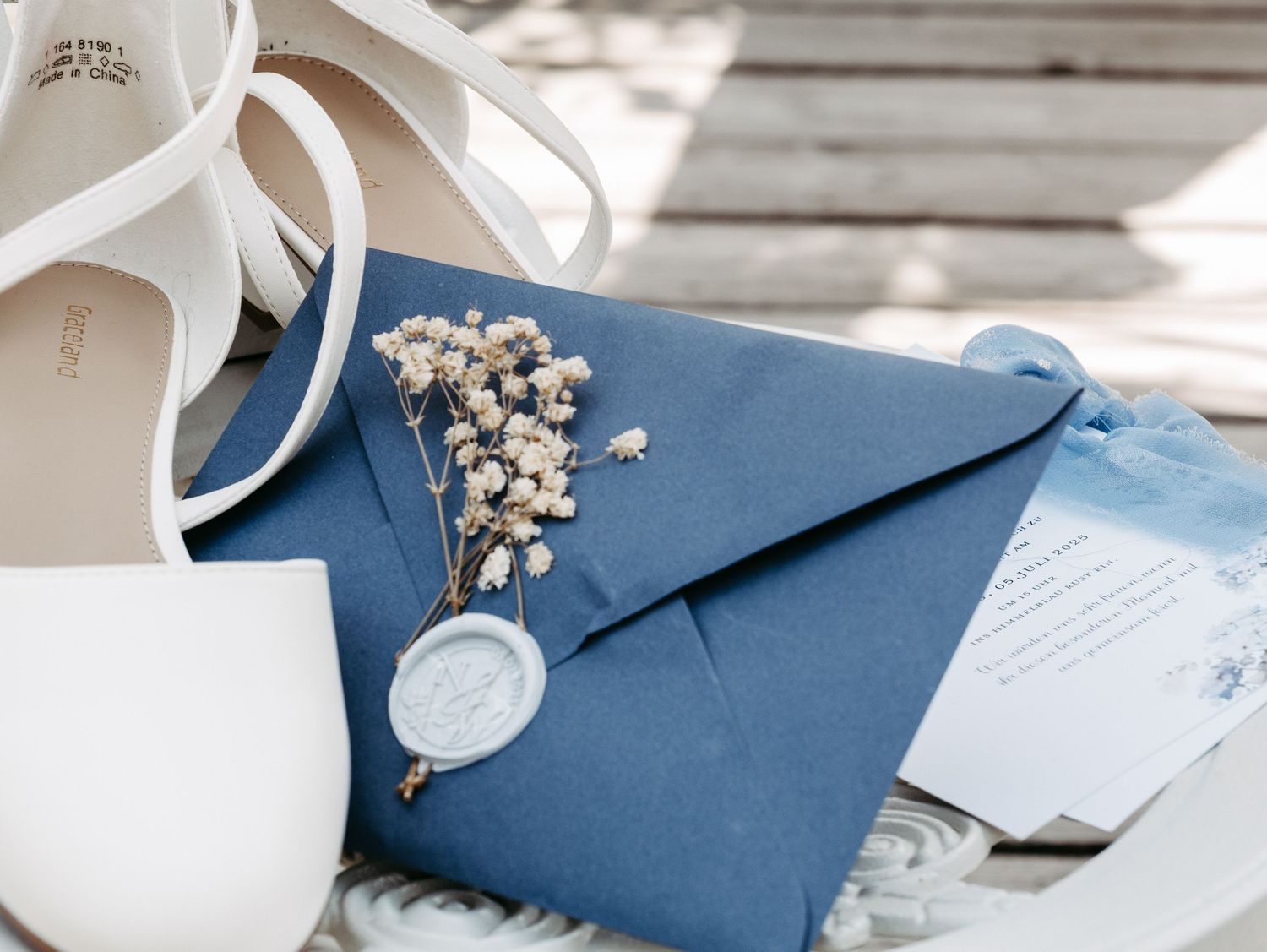 Blue envelope with wax seal, dried baby's breath flowers, and white wedding shoes on a wicker surface.