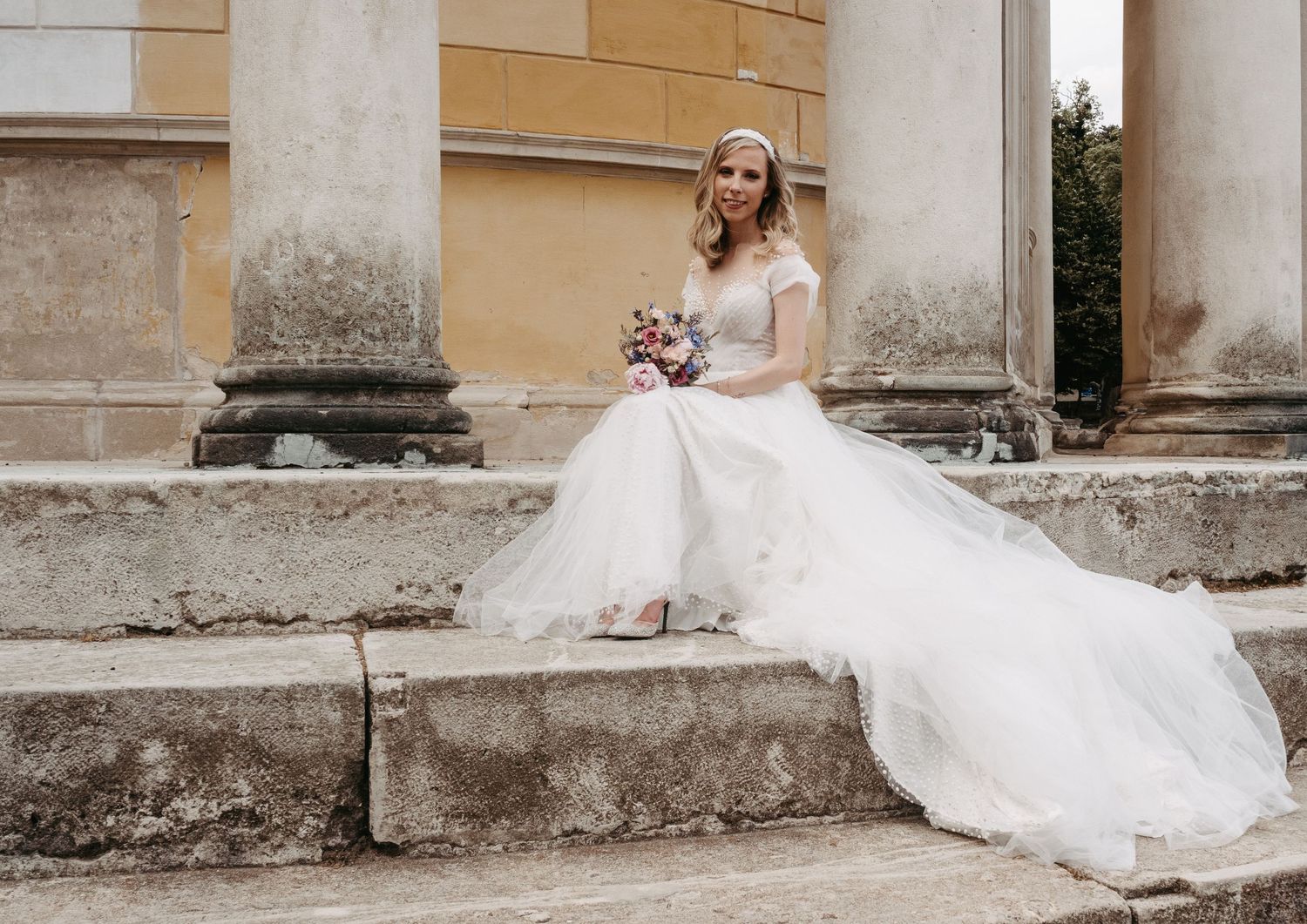 Bride in white off-shoulder gown sits on stone steps holding bouquet, with classical columns in background.