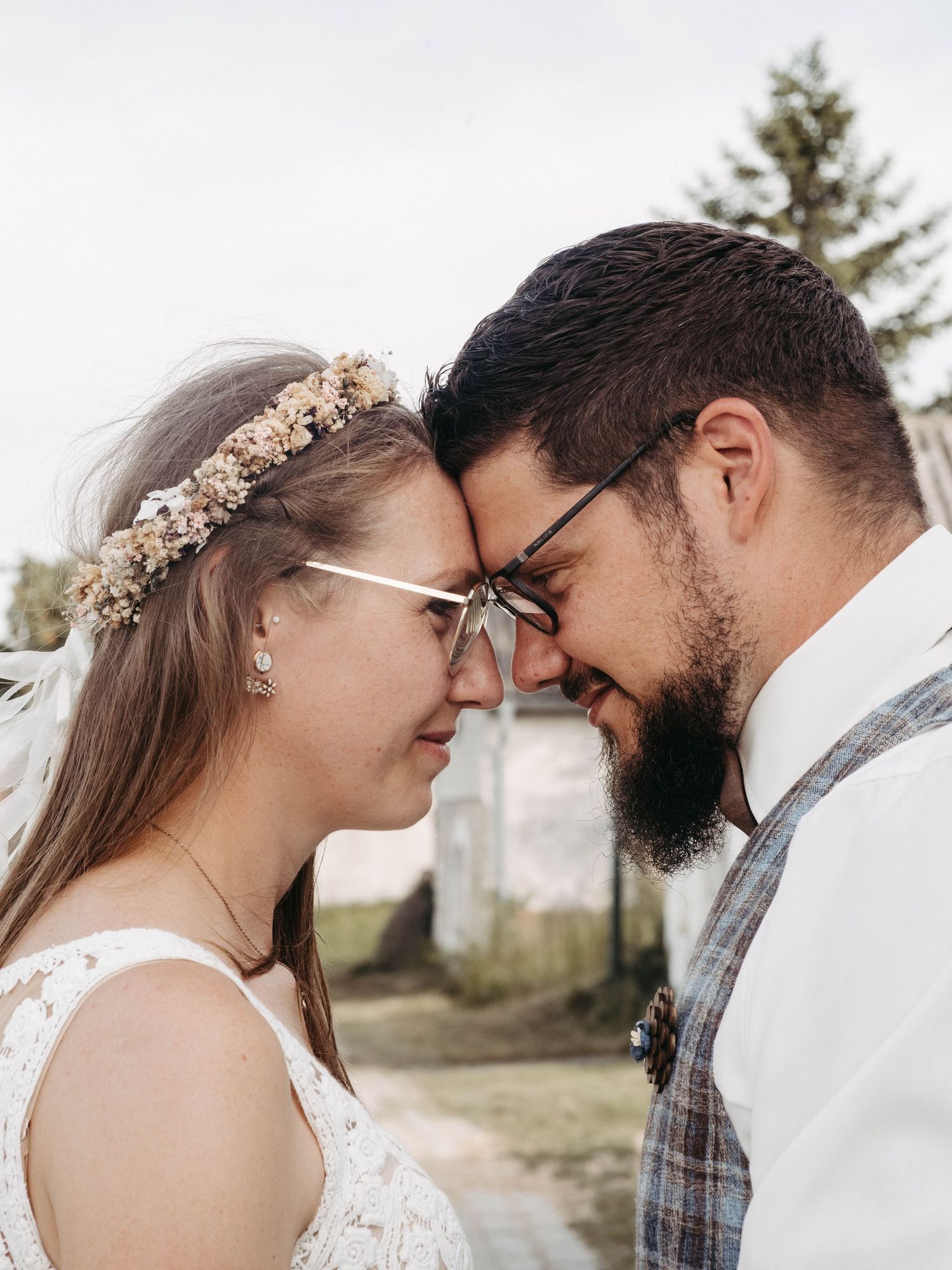 A bride and groom touch foreheads and smile on their wedding day, bride wearing floral crown and groom sporting a beard.