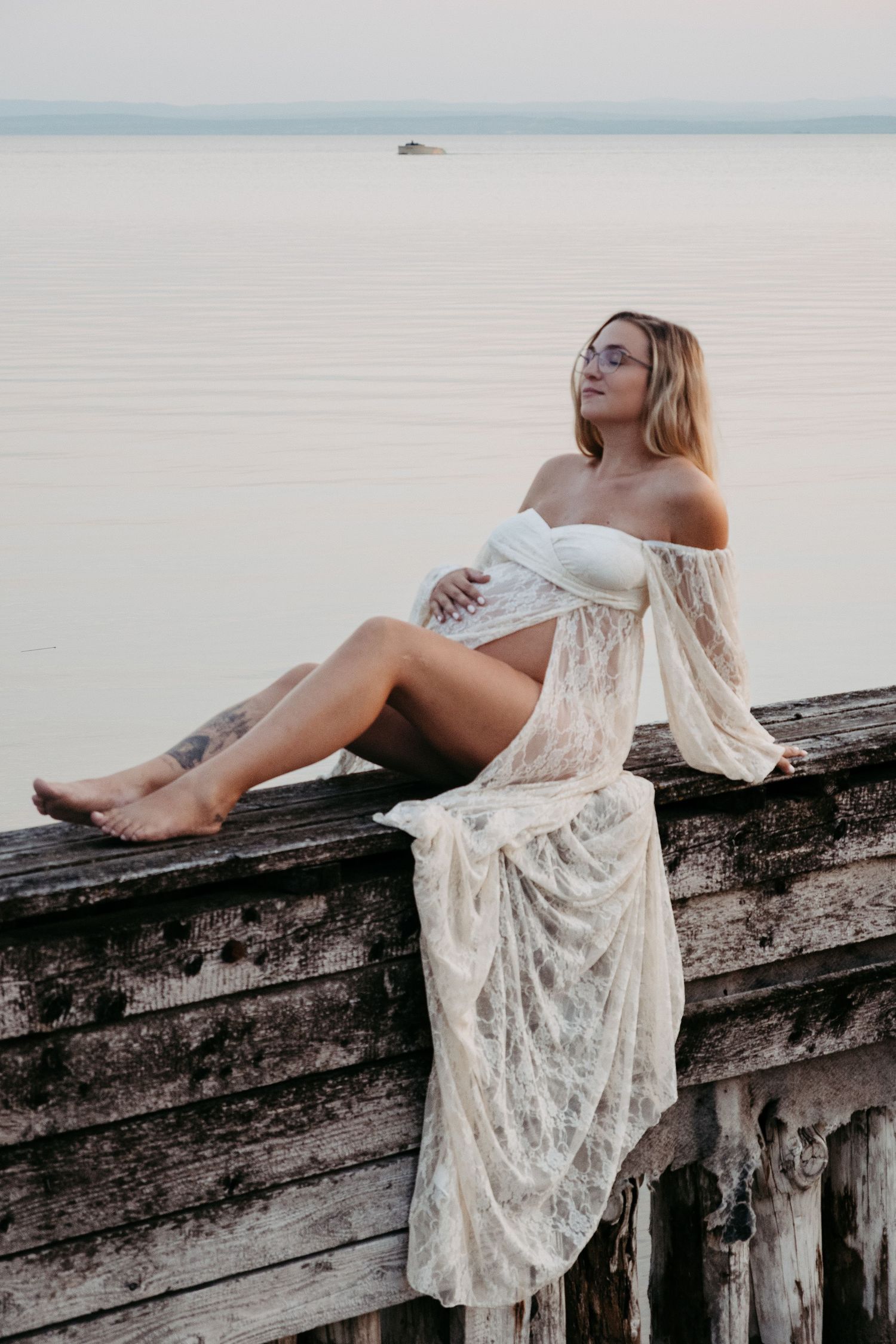 Artistic portrait of woman in white lace dress posing on weathered wooden pier at dusk.