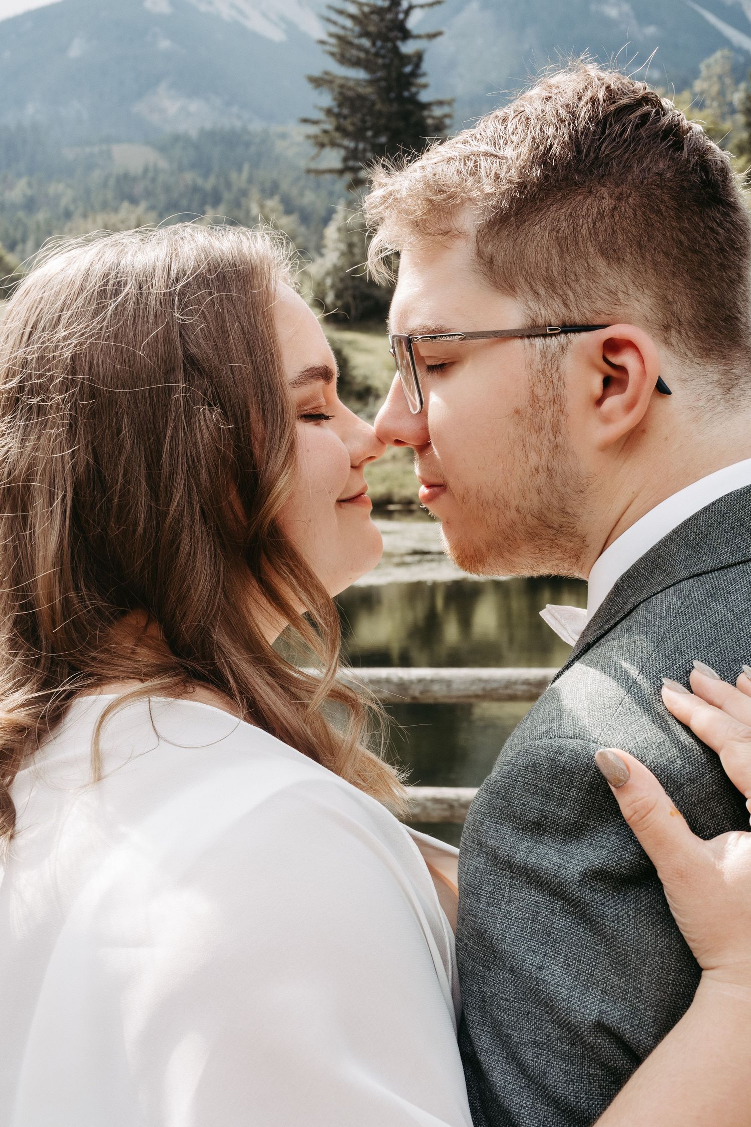A couple shares a romantic almost-kiss moment outdoors with mountain scenery and a pond in the background.