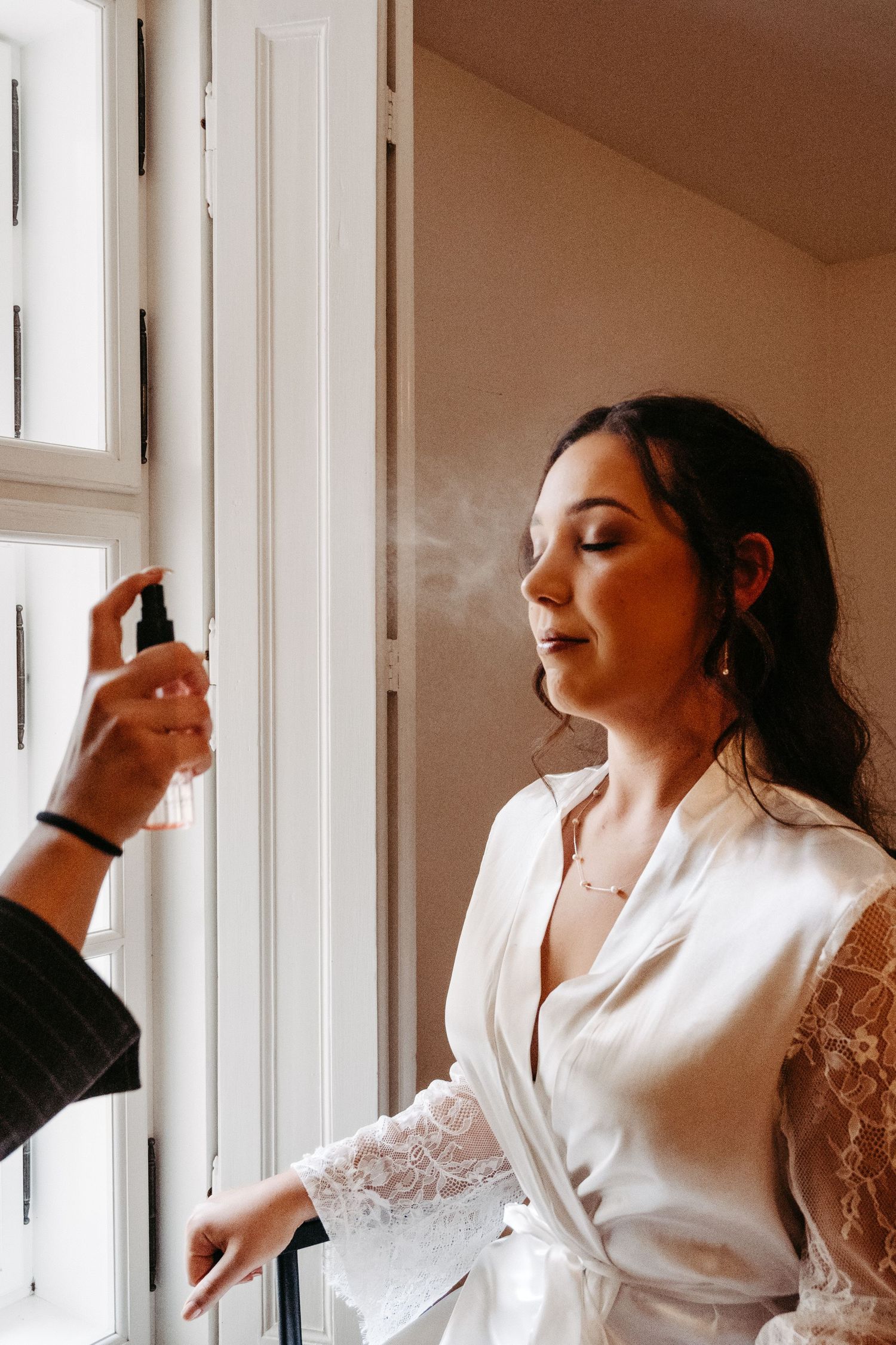 Bride in white lace robe getting hair sprayed near window during wedding preparations.