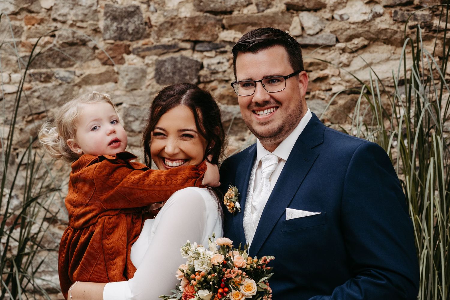 Smiling bride and groom posing with young child in rust dress at wedding.