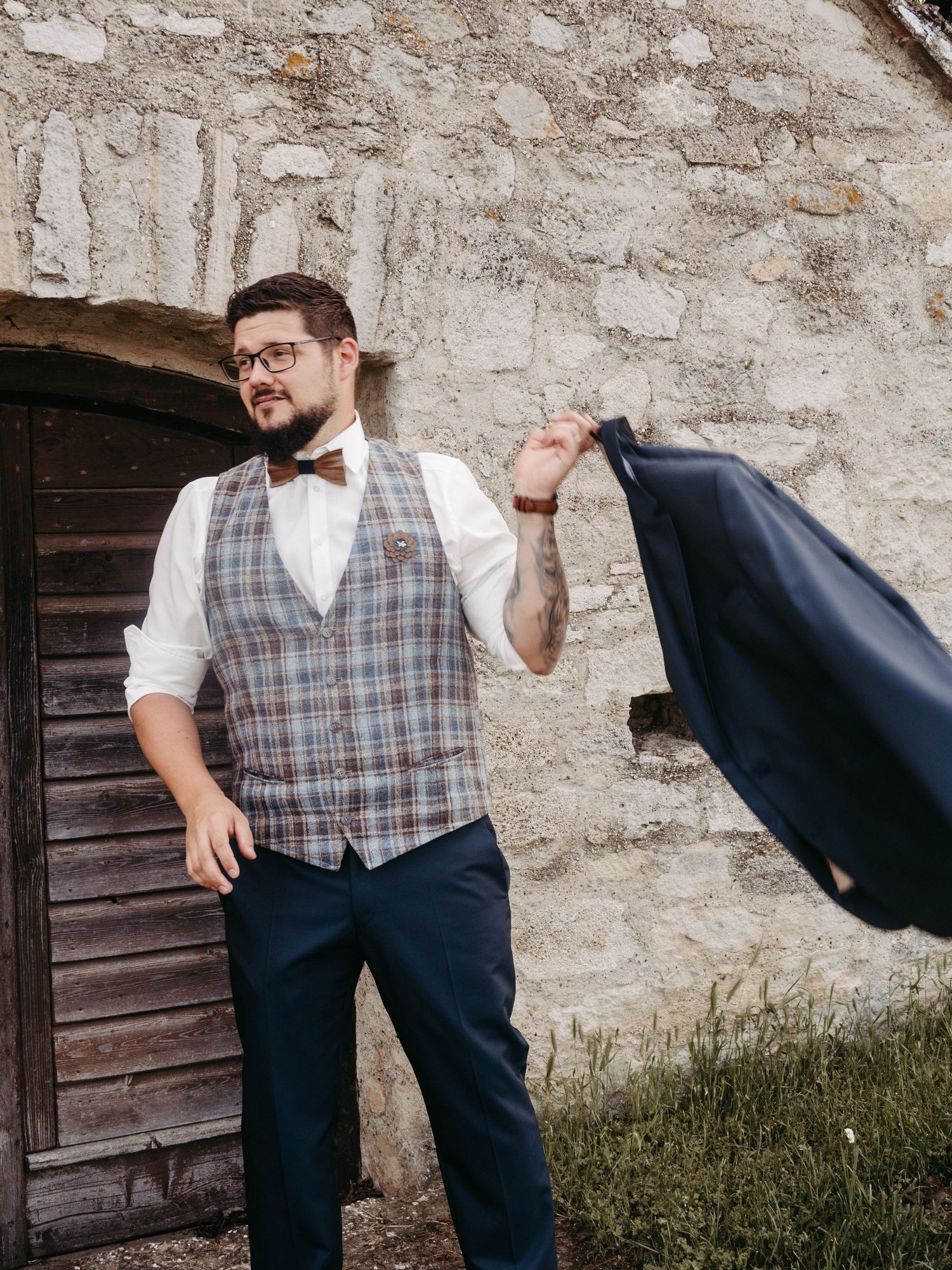 Stylish groom in plaid vest, bow tie, and navy trousers holding jacket outside rustic stone wall.