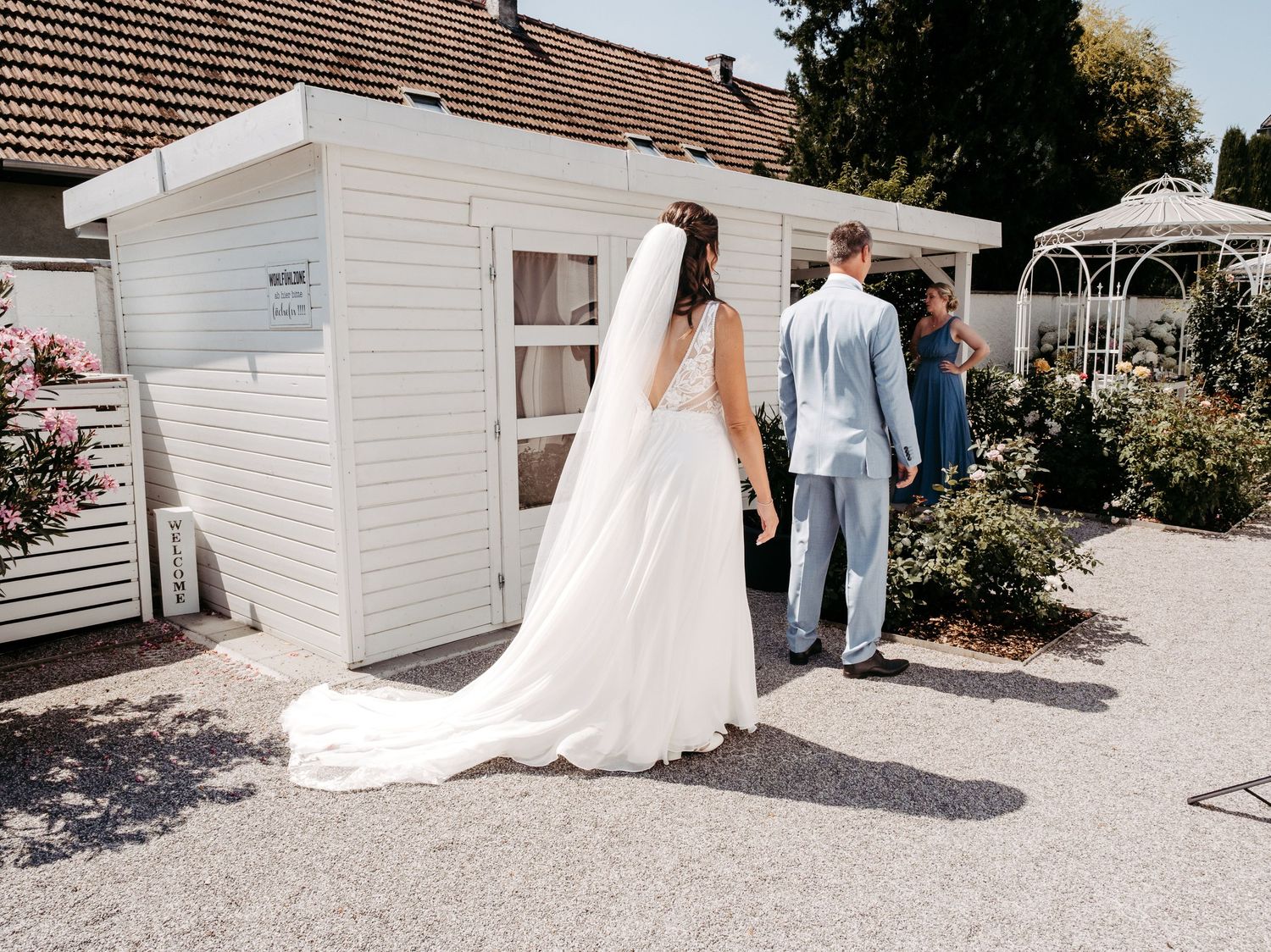 Bride in white gown and veil walks with groom in light blue suit toward white building at outdoor wedding venue.