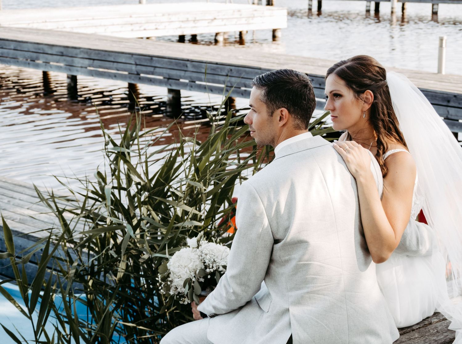 Bride and groom sitting together by waterfront, bride in white veil gazing at groom in light suit.