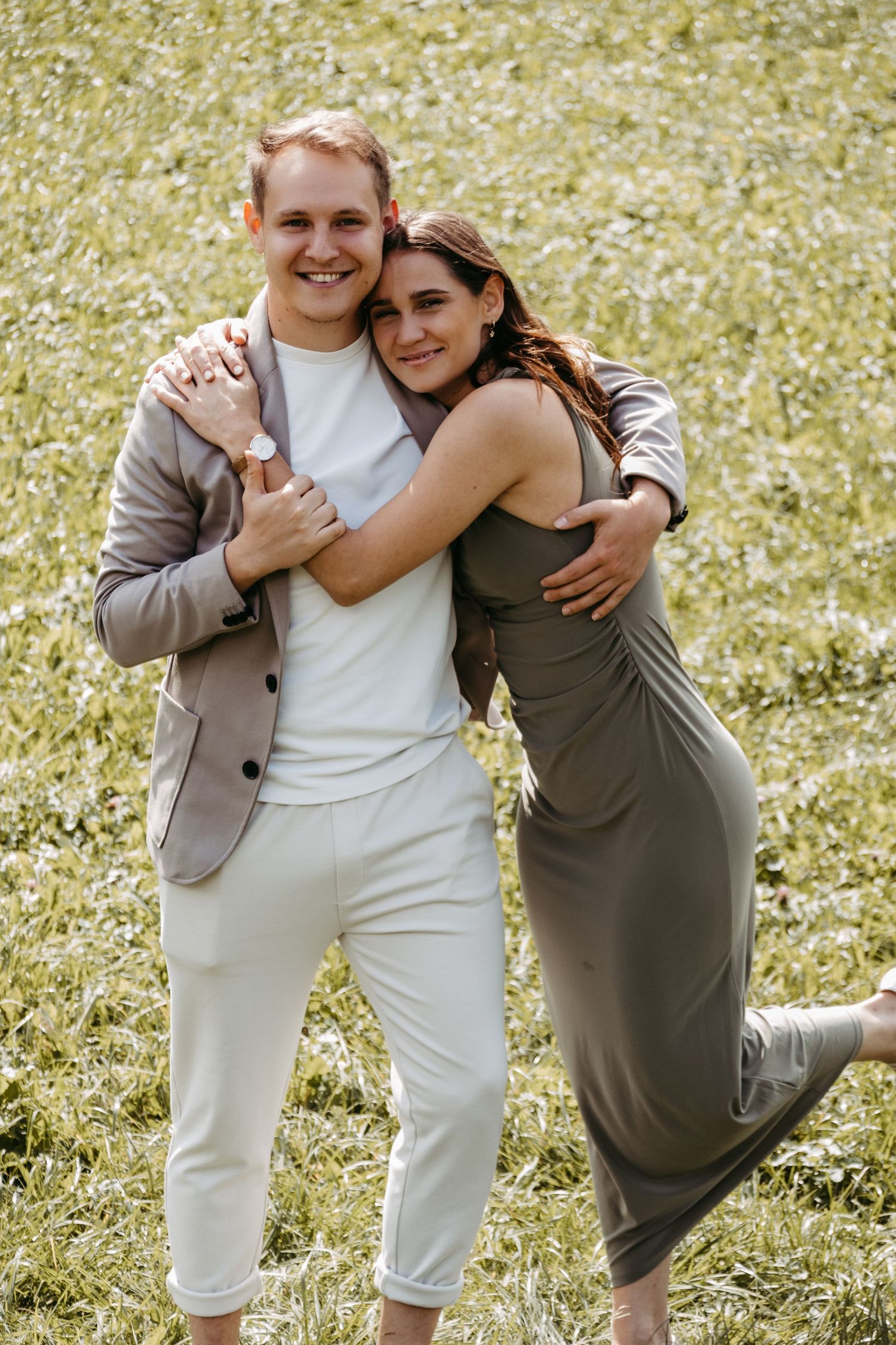 Happy couple embracing outdoors on green grass, woman in olive dress hugging smiling man in blazer and white pants.