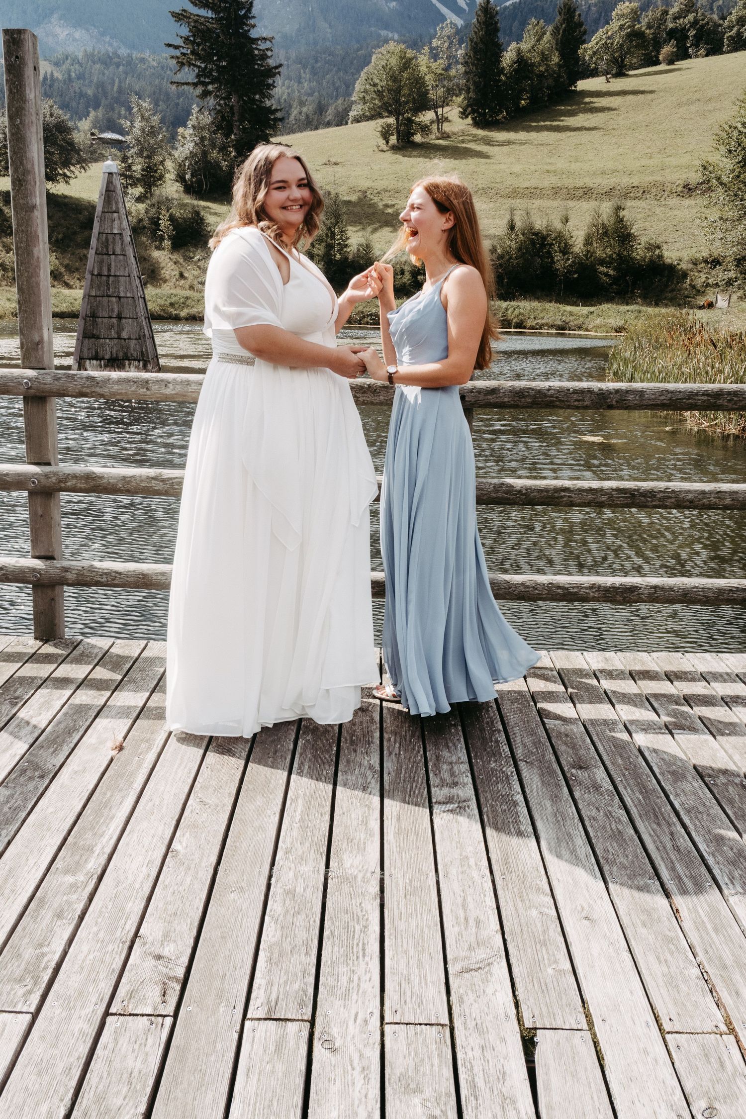 Bride in white gown holds hands with bridesmaid in blue dress on wooden dock by mountain lake.