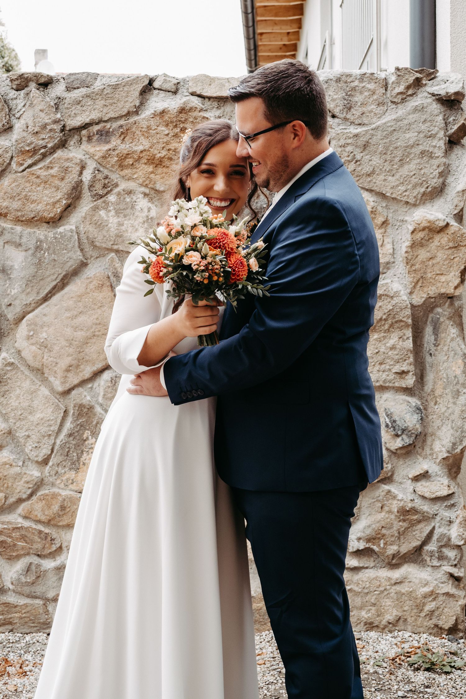 A groom in a navy suit embraces his bride holding an orange and white bouquet against a stone wall.