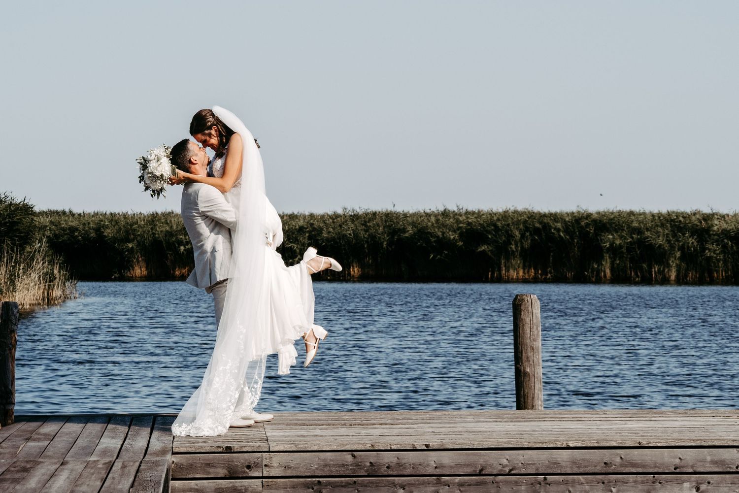 Groom lifting bride in white gown on wooden dock by lake, sharing romantic kiss on sunny wedding day.