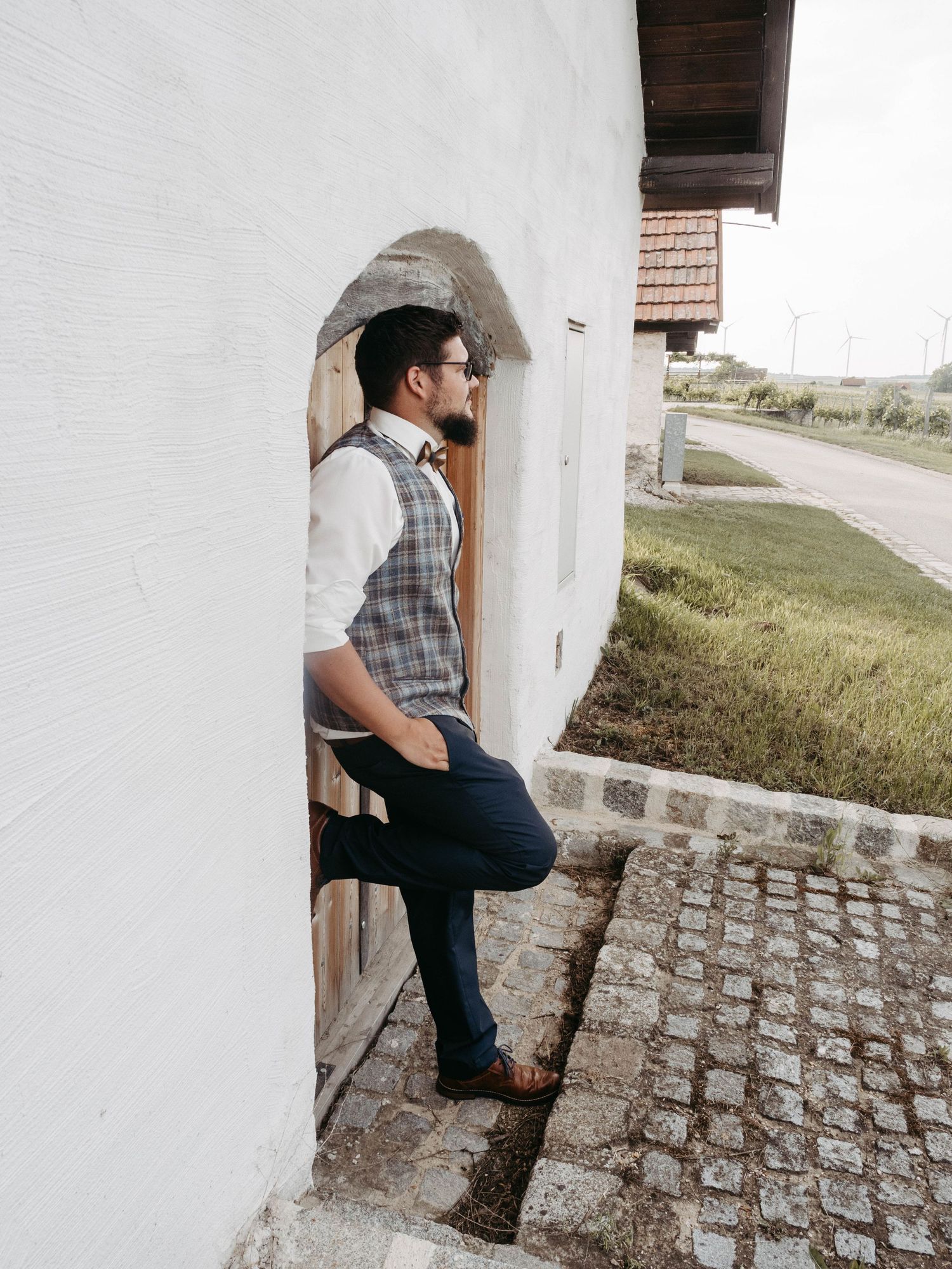 Man in plaid vest and navy trousers leaning against a white stone archway doorway on a cobblestone path.
