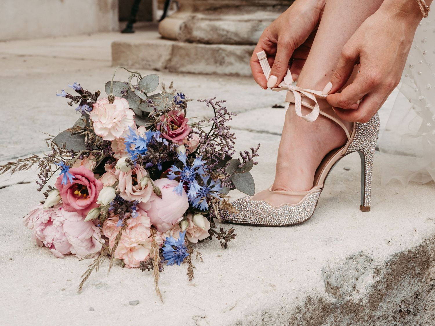 Bride tying glittery heels on stone steps beside a lush bouquet of pink peonies and blue thistle flowers.