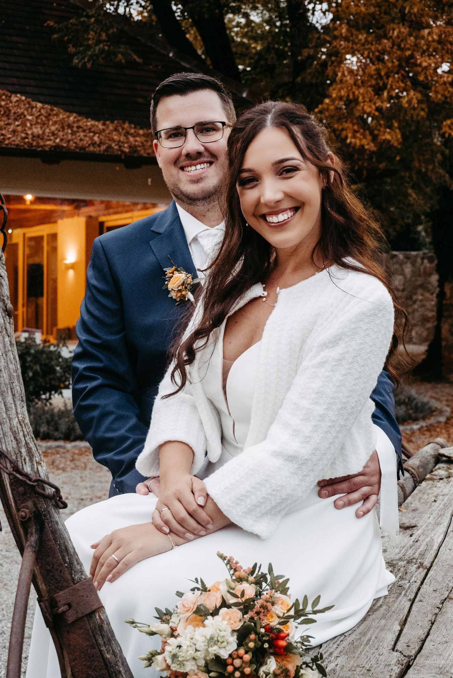 Romantic autumn wedding photos featuring a couple in white dress and navy suit against warm fall foliage and glowing lights.