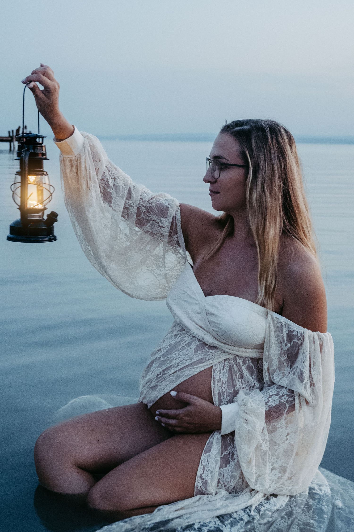 Artistic photos with antique lantern in white lace dress at ocean's edge during blue hour.