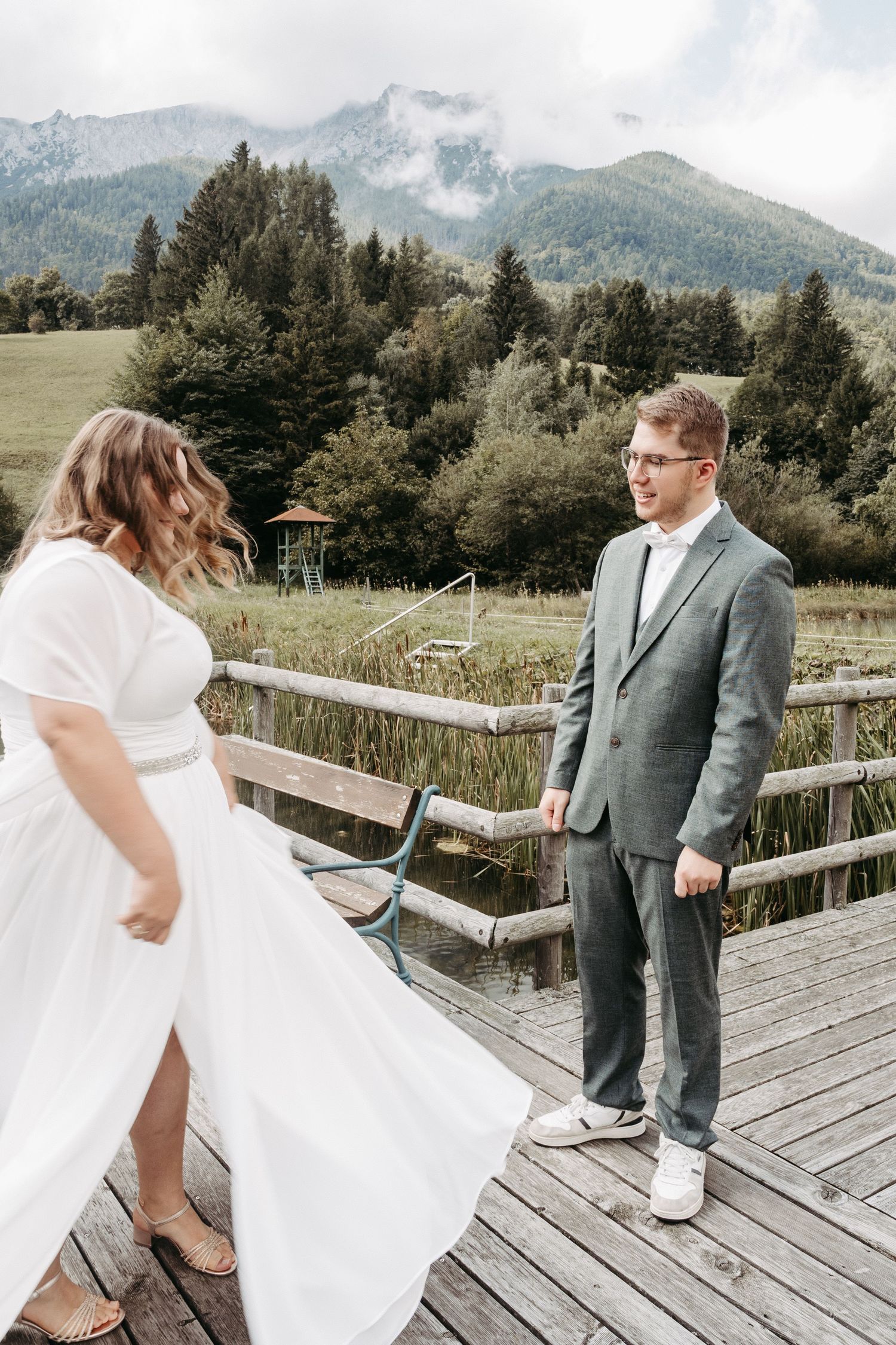 Bride in white dress and groom in gray suit stand on wooden dock with mountain backdrop.