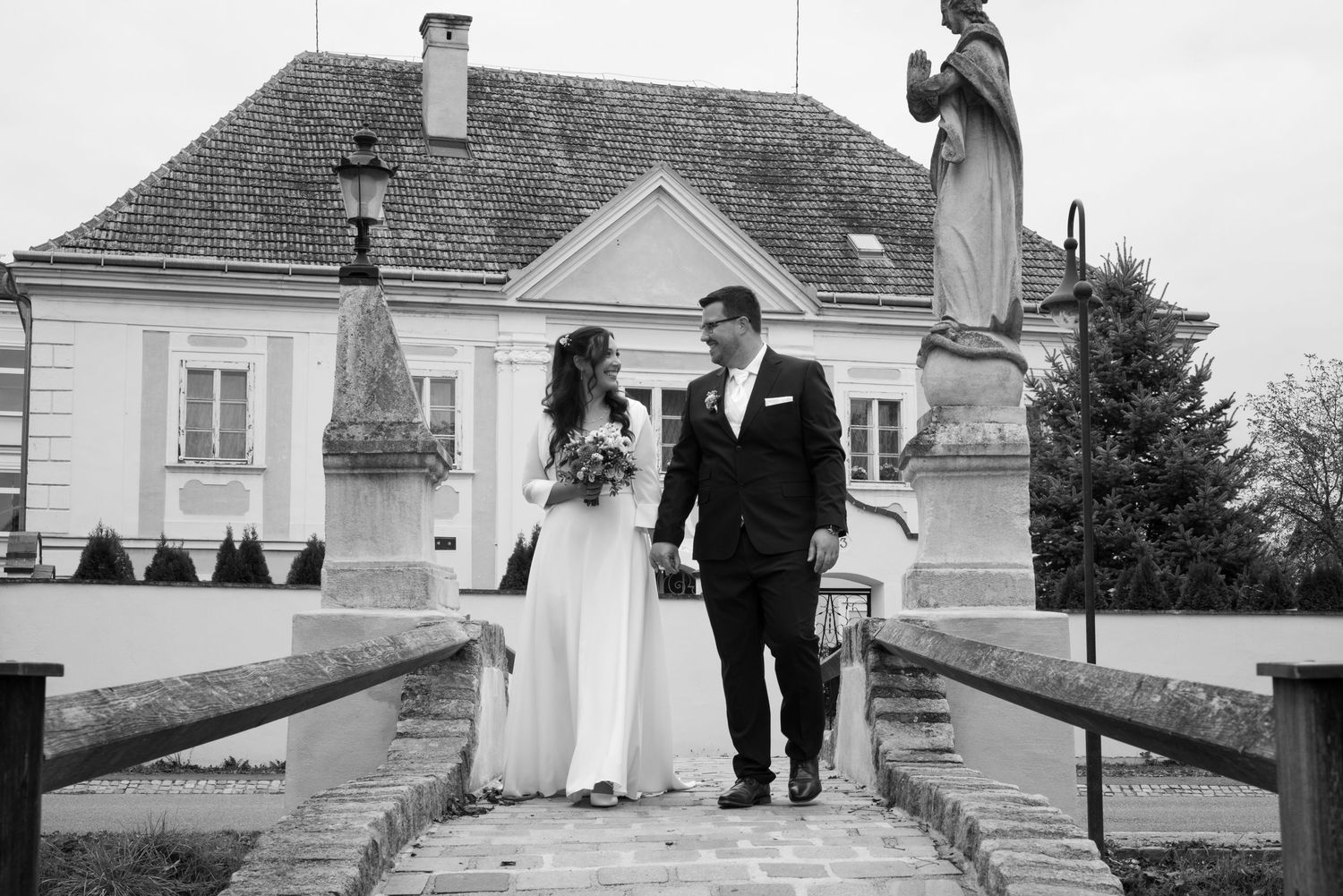 Black and white wedding photo of bride and groom walking hand in hand on steps before a historic European manor.