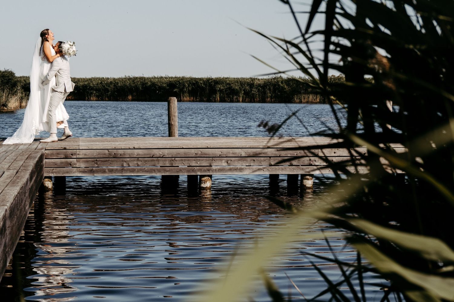 Bride and groom stand on a wooden dock overlooking a serene lake surrounded by lush green trees on a sunny day.