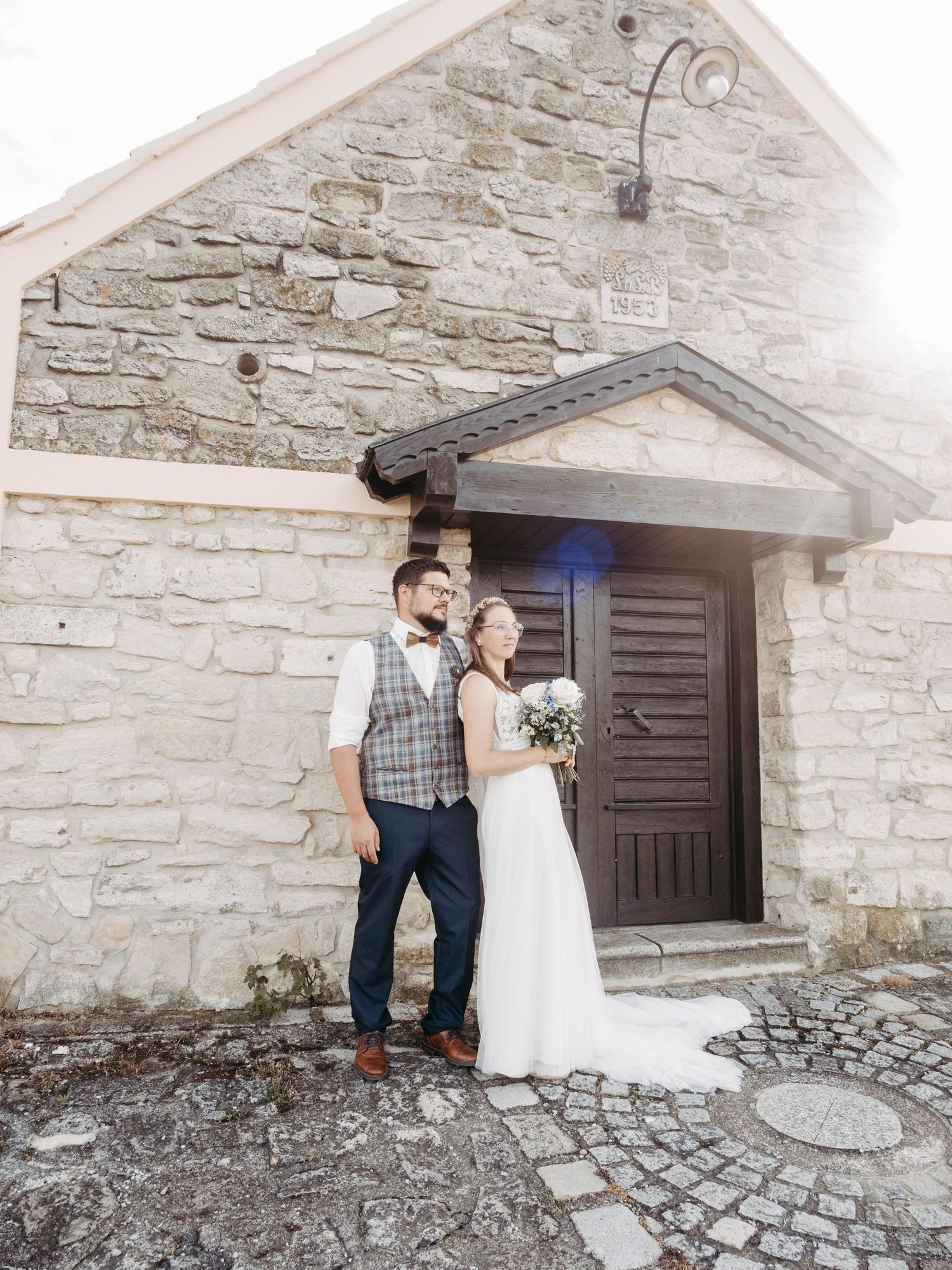 Bride and groom posing in front of a rustic stone chapel, bride holding white bouquet, groom in plaid vest.