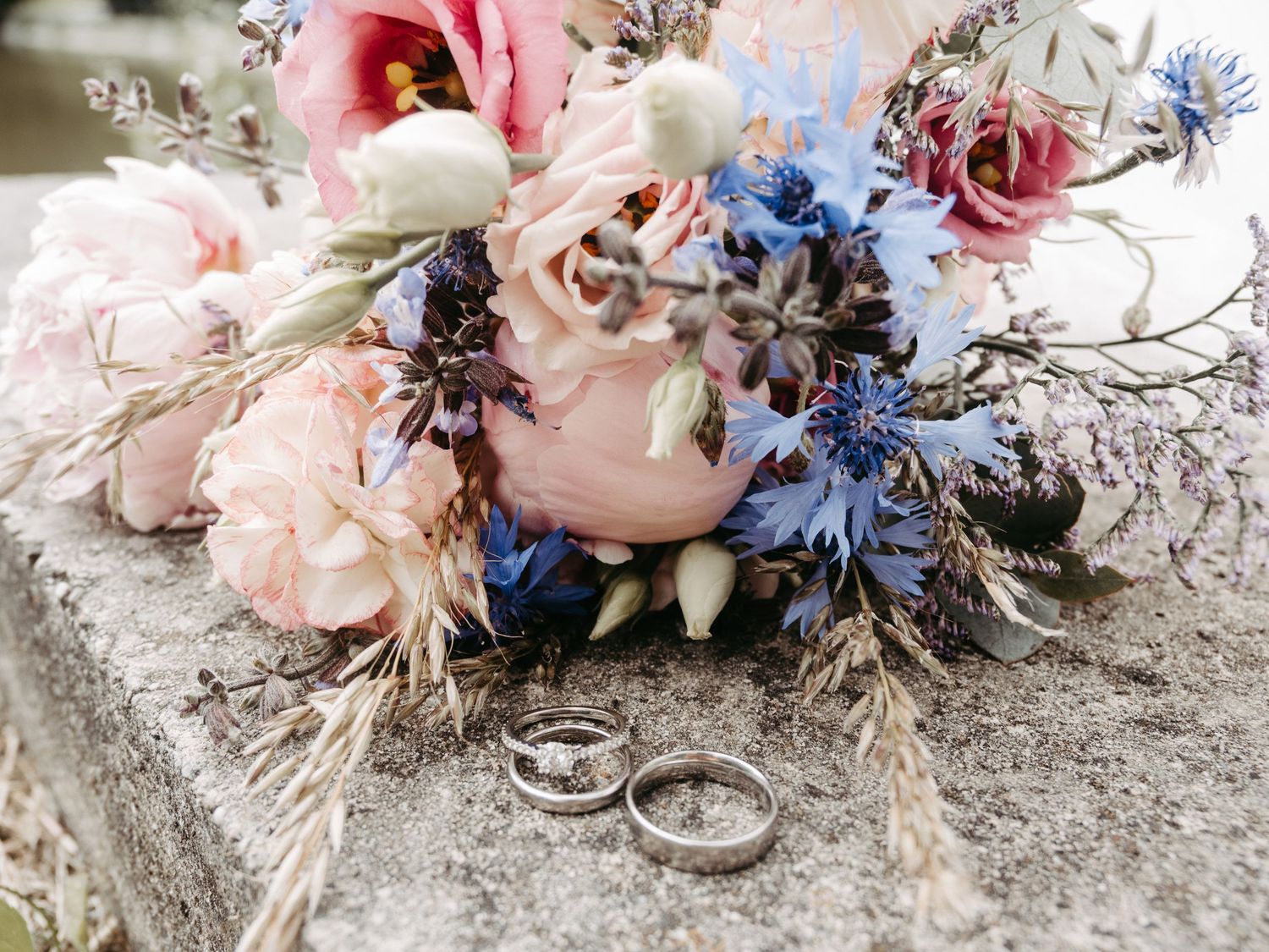 Wedding rings resting beside a romantic bouquet of pink roses and blue cornflowers on a stone surface.