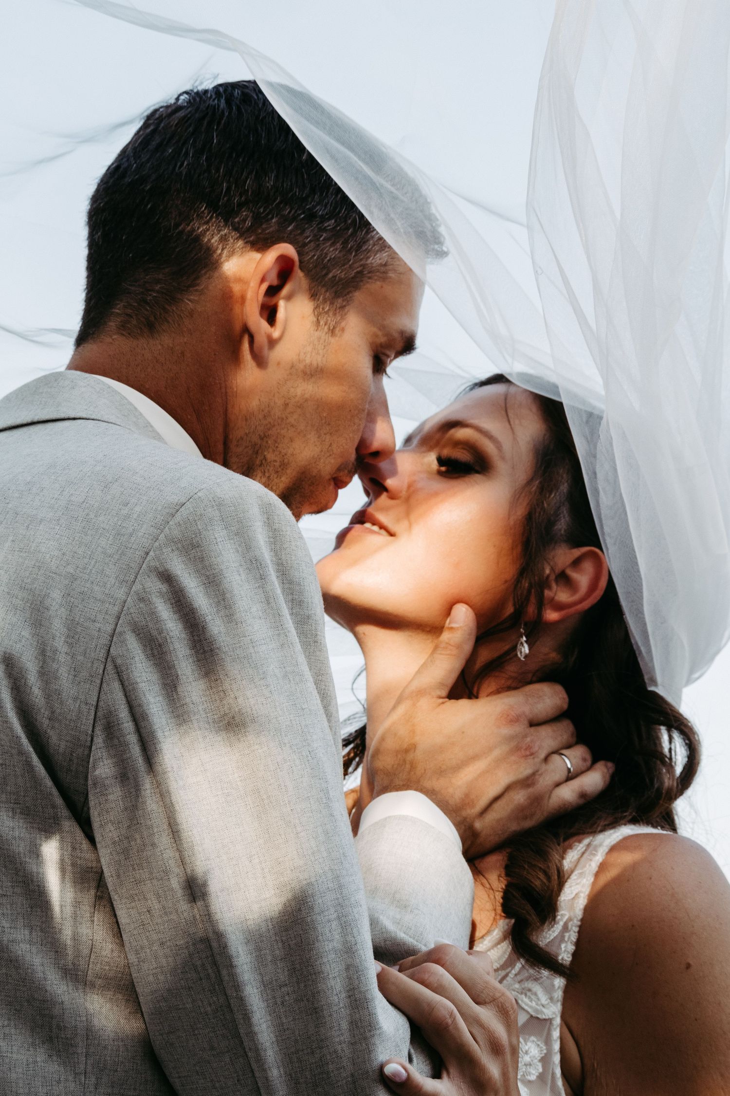 Bride and groom share an intimate moment under a flowing white veil, with groom tenderly holding bride's face.