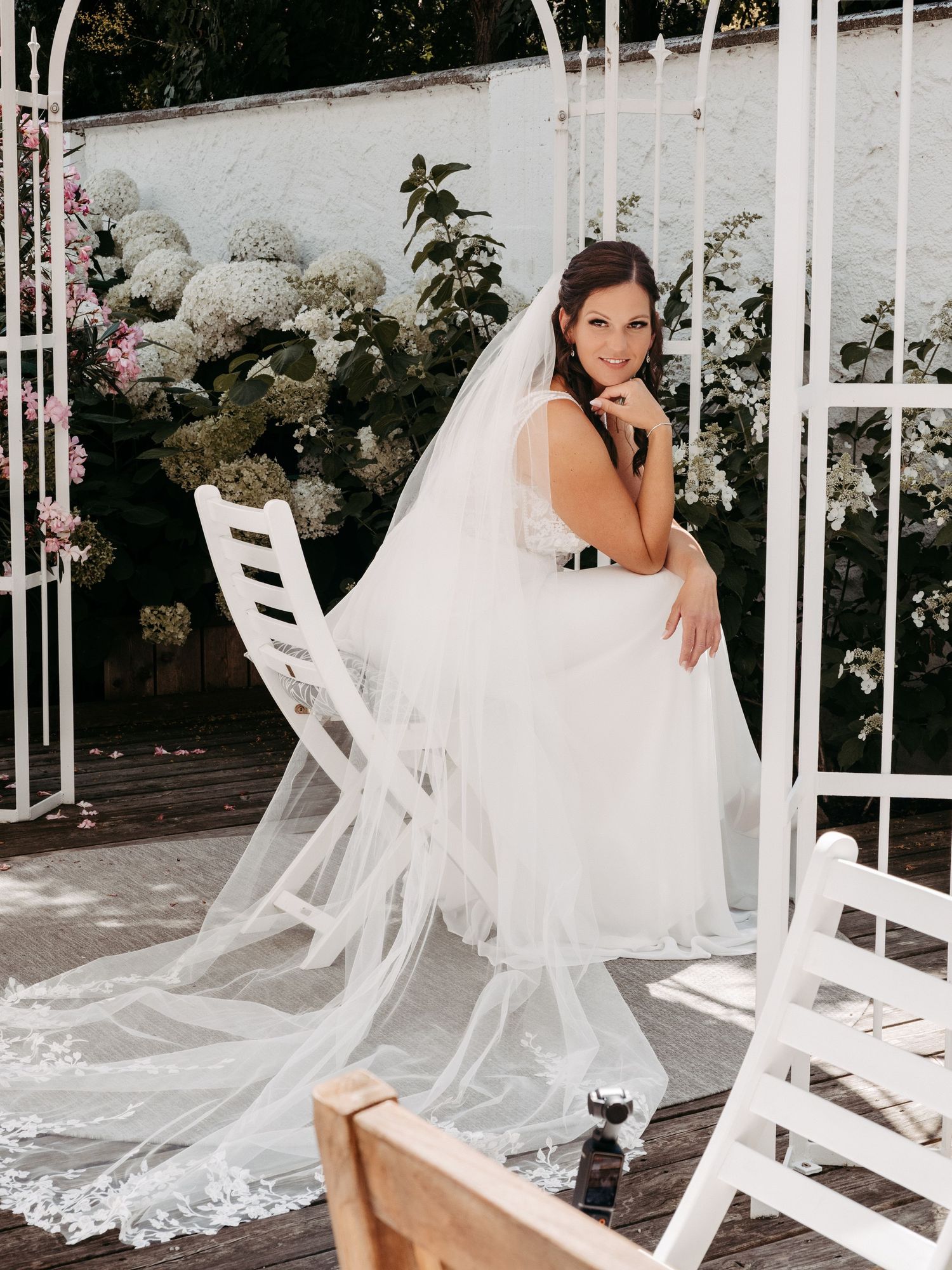 Bride in white wedding gown and long lace veil seated on white chair surrounded by blooming hydrangeas outdoors.