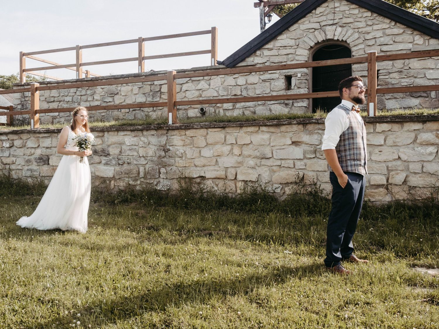 Bride in white gown approaches groom in plaid vest during first look at rustic stone barn wedding venue.