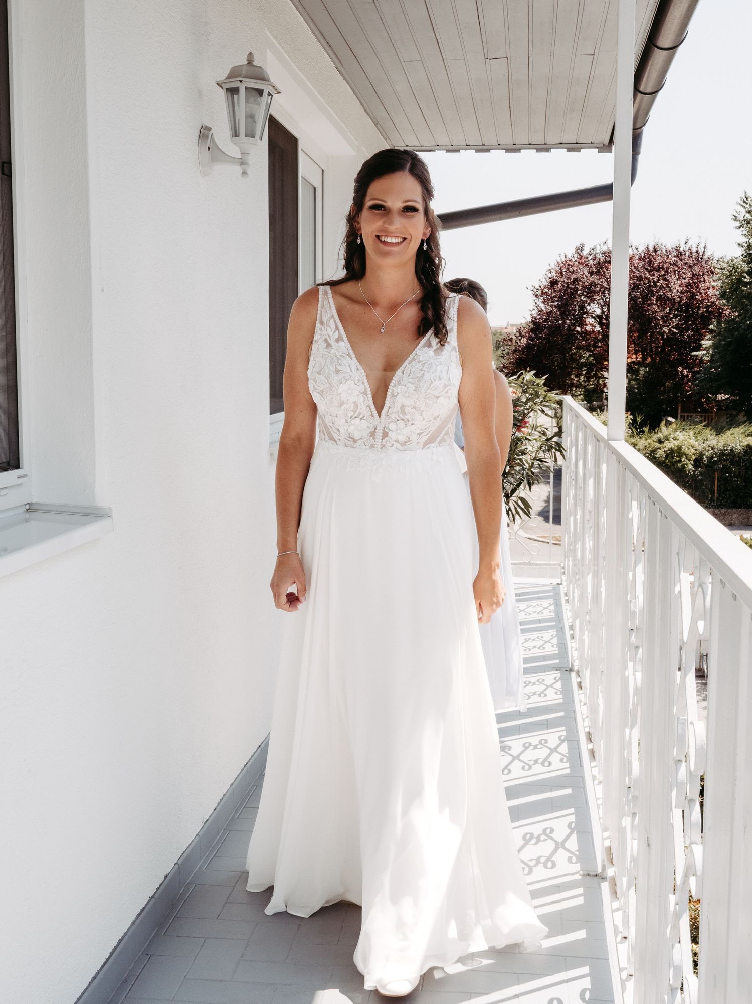 Smiling bride in a white V-neck lace wedding gown standing on a sunlit balcony with greenery in the background.