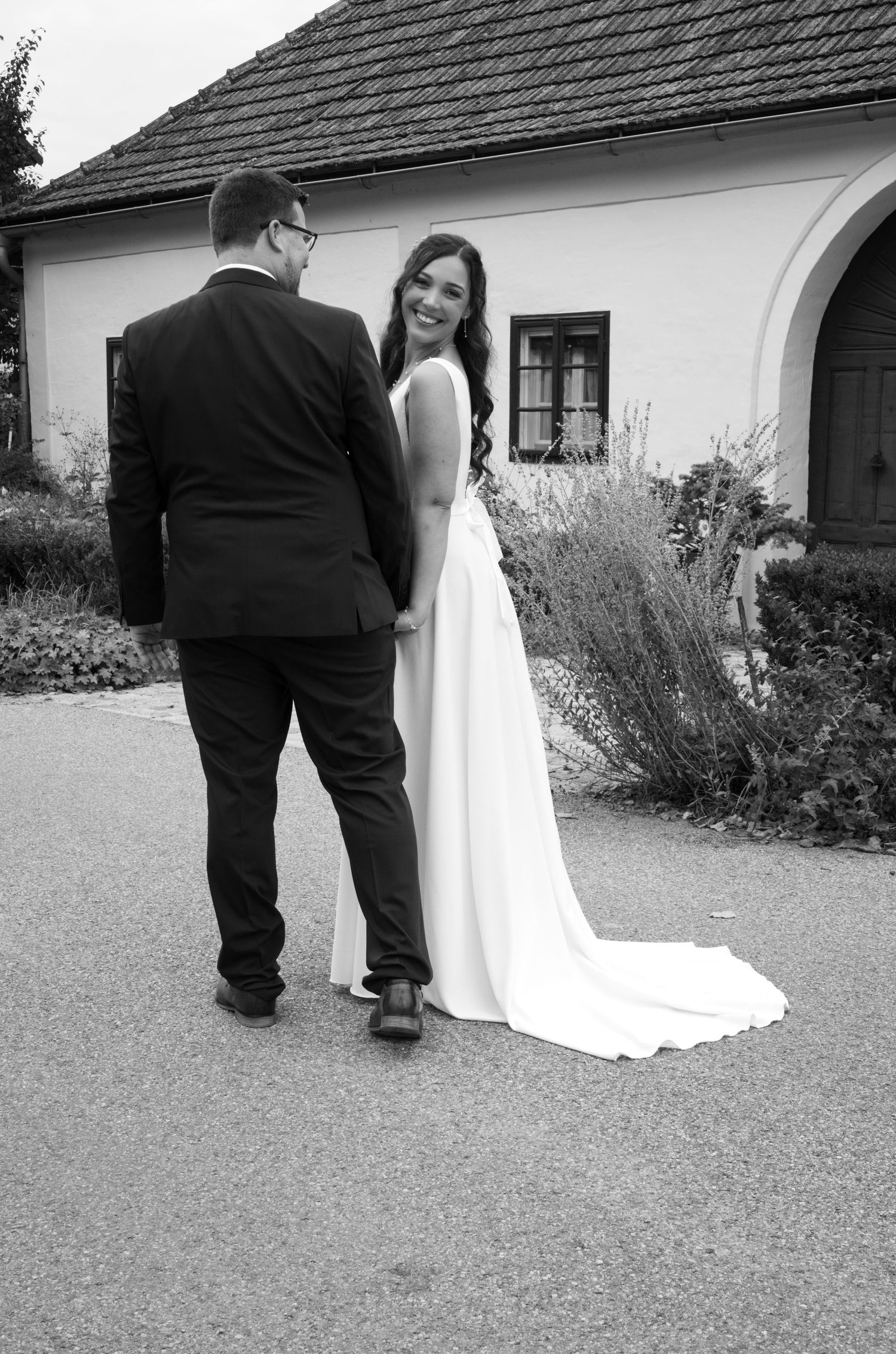 Black and white photo of smiling bride in flowing gown walking with groom outside.