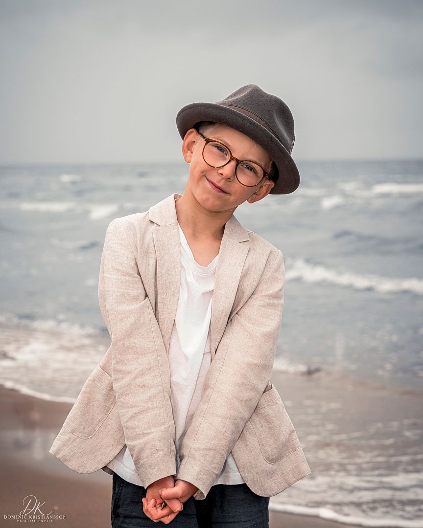 Young boy in gray hat, glasses, and beige blazer poses confidently on a sandy beach with ocean waves behind him.