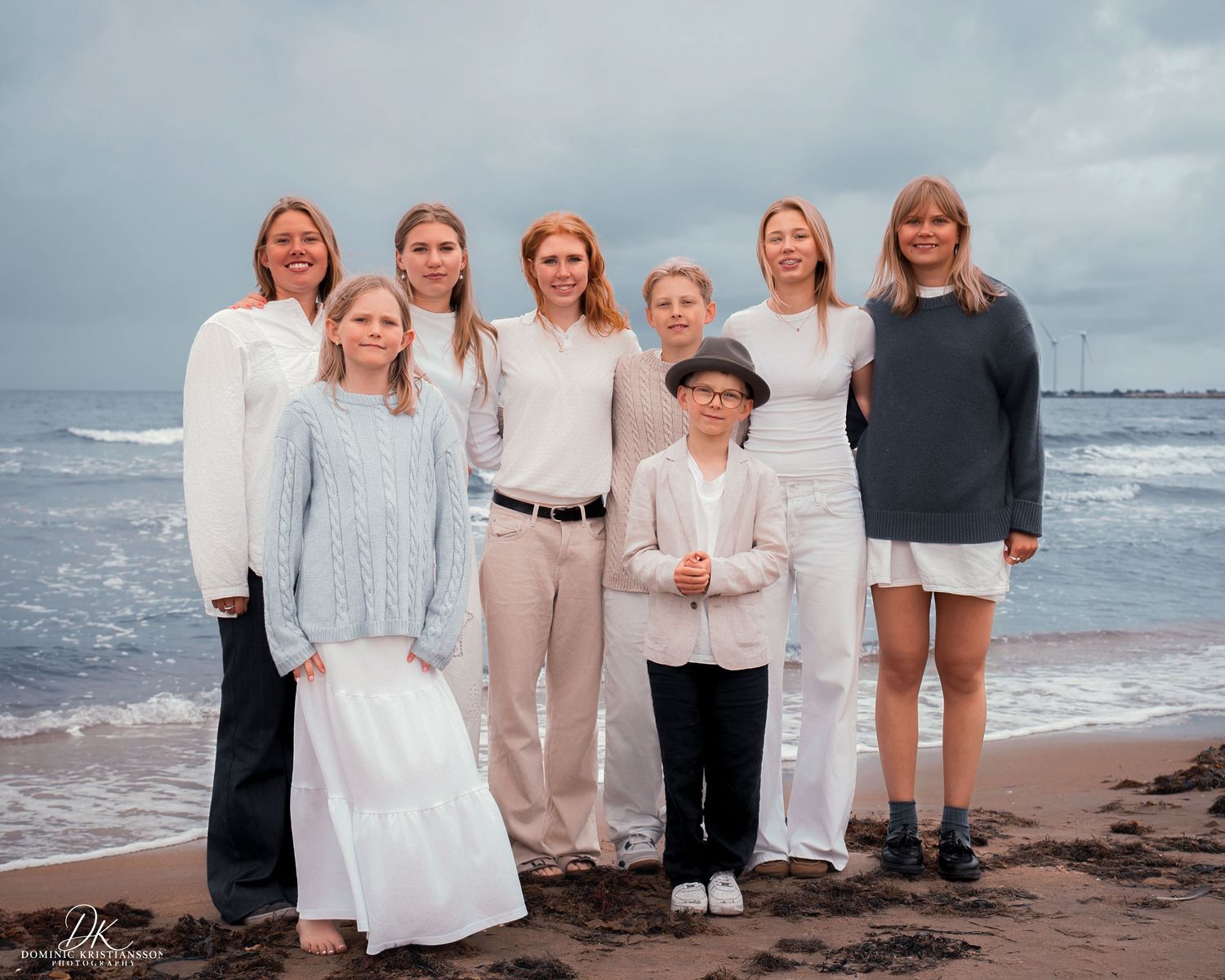 Group of eight women and girls posing together on a sandy beach with ocean waves in the background.
