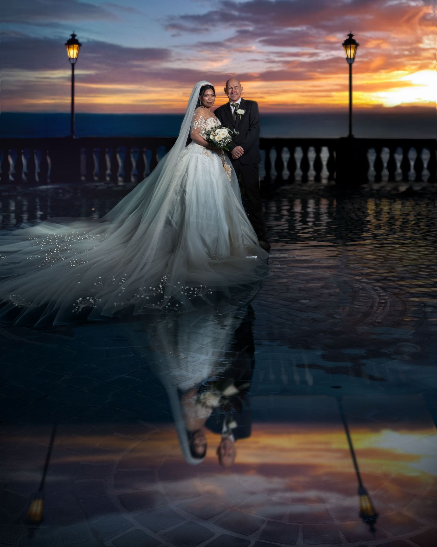 A wedding couple poses on a balcony at sunset with glowing lampposts and their reflection in wet pavement.