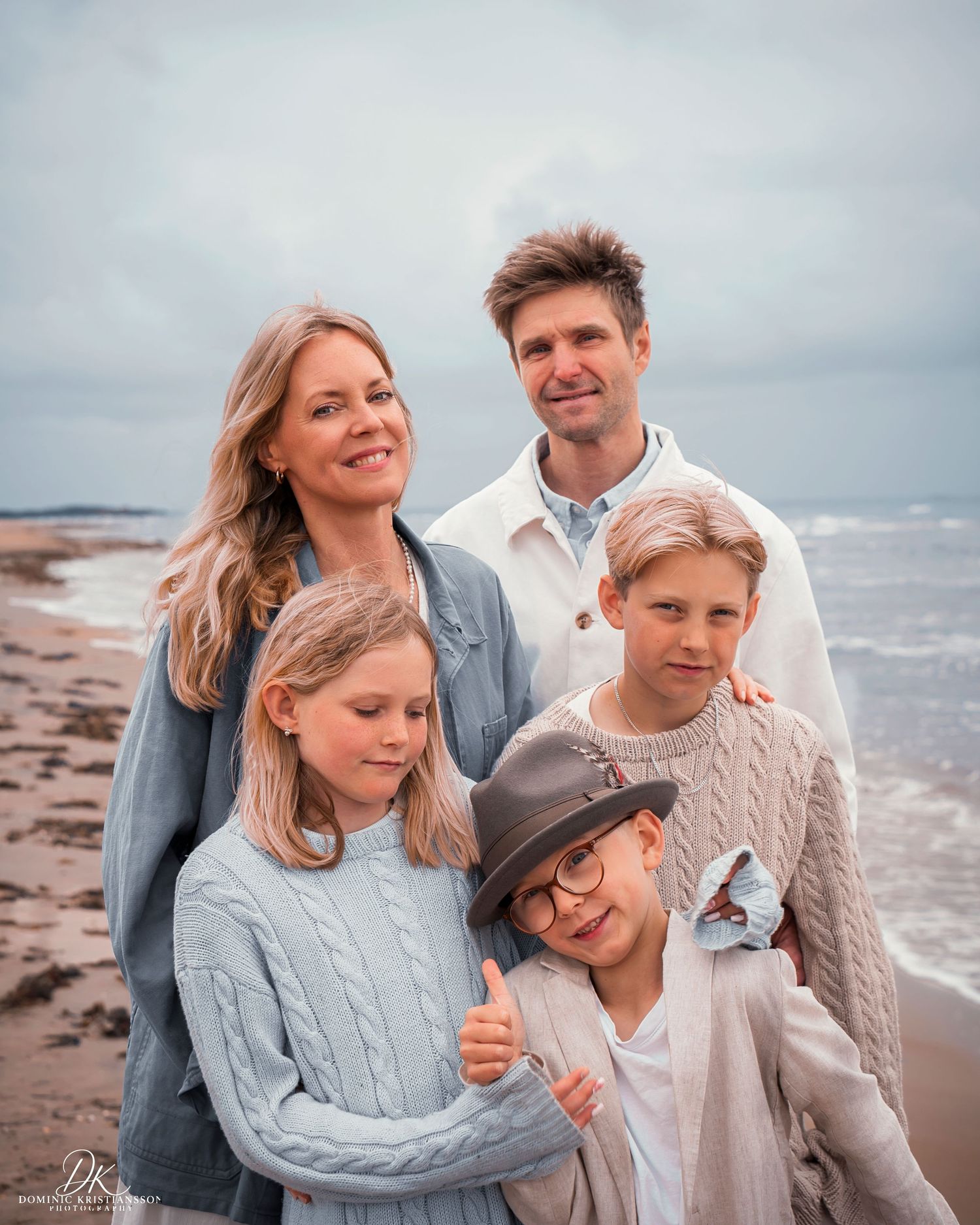 A family of five poses together on a beach, wearing cozy knit sweaters, with the ocean visible behind them.