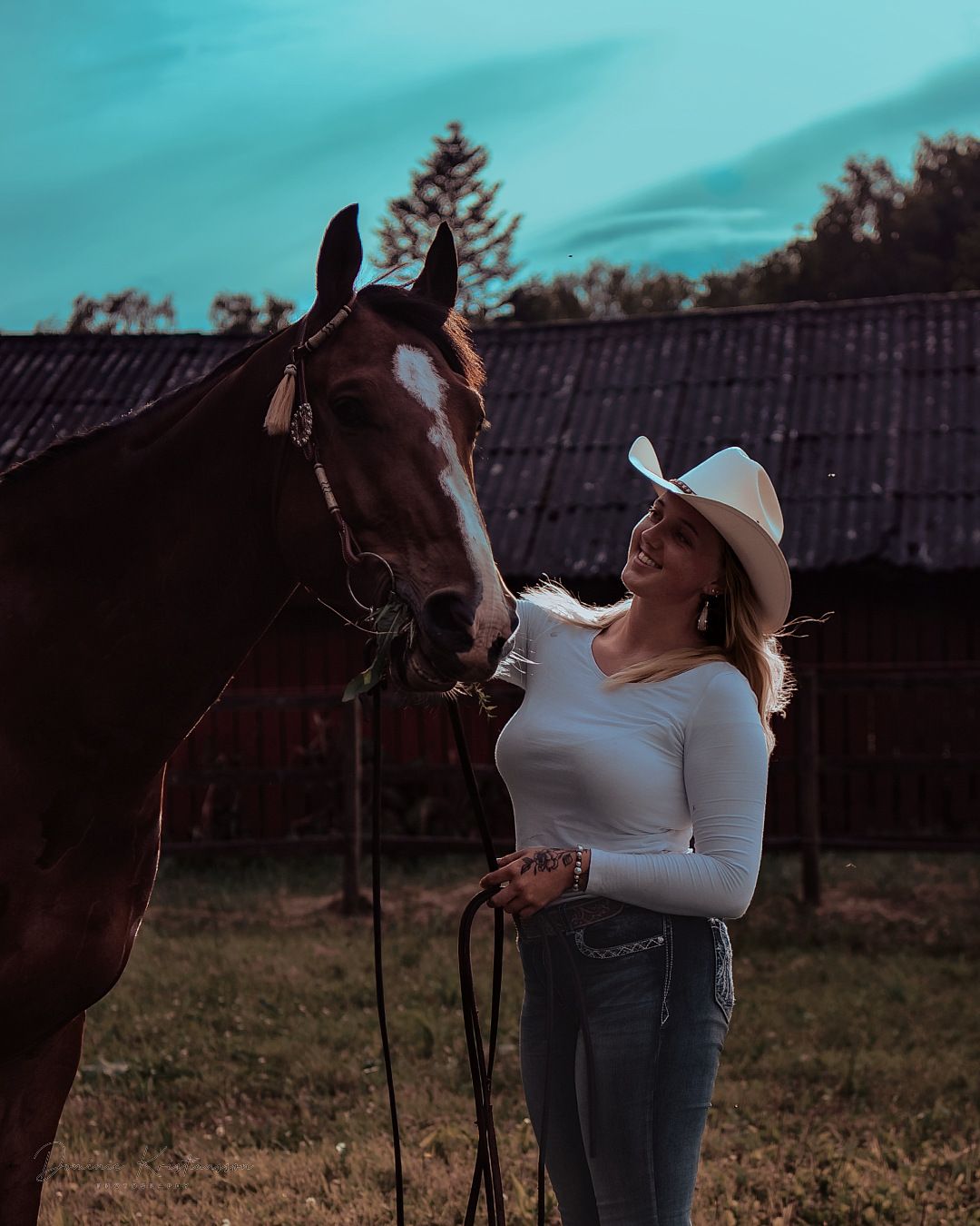 A bay horse and rider in white cowboy hat share a moment at dusk against a rustic barn backdrop.