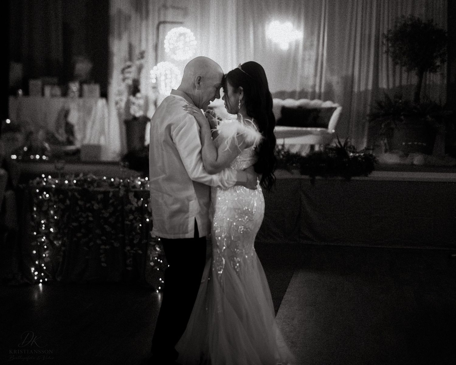 A couple shares a romantic dance at a dimly lit wedding reception with twinkle lights in the background.