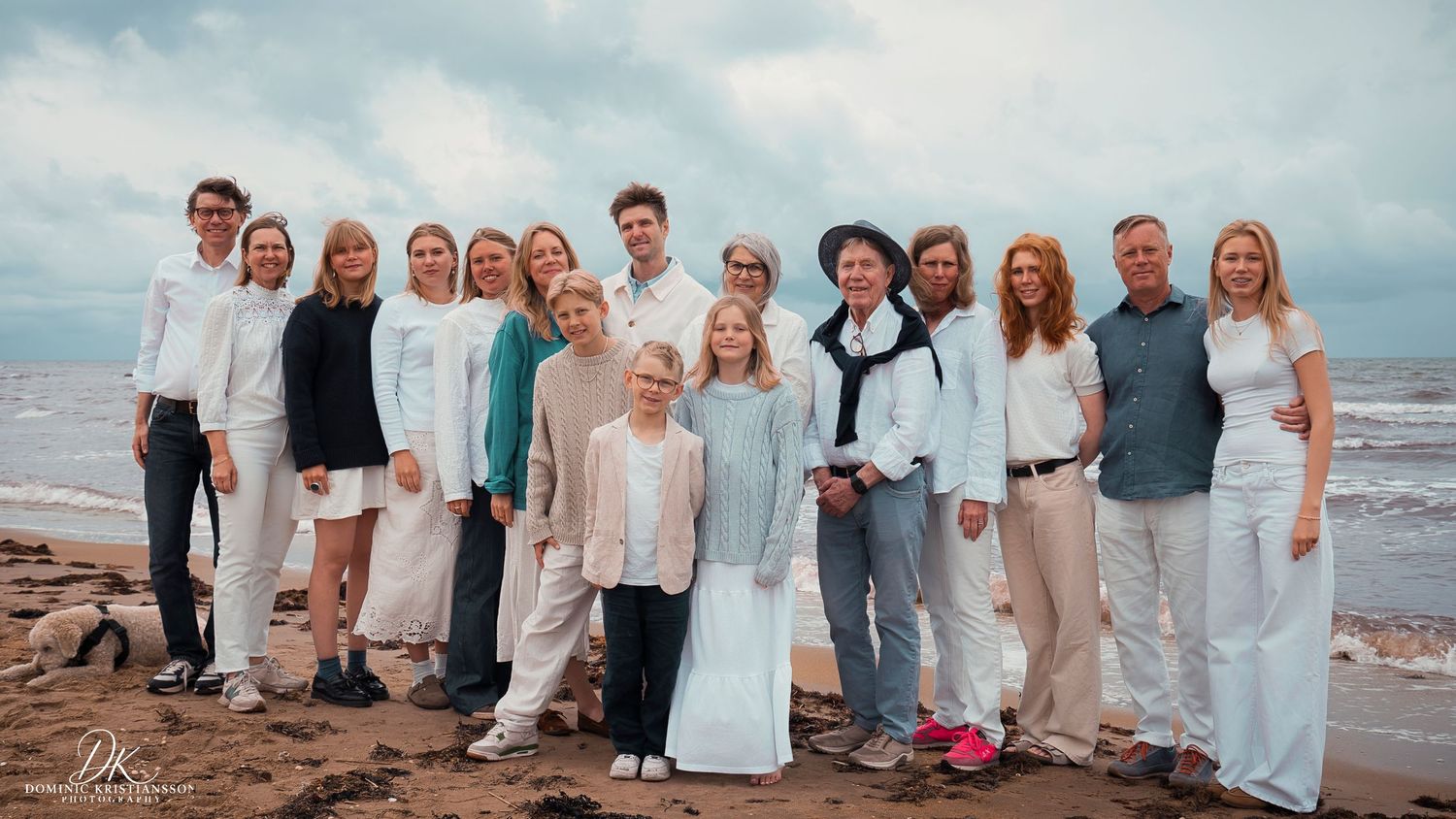 Large family group of about fifteen people posing together on a beach with cloudy skies and ocean behind them.