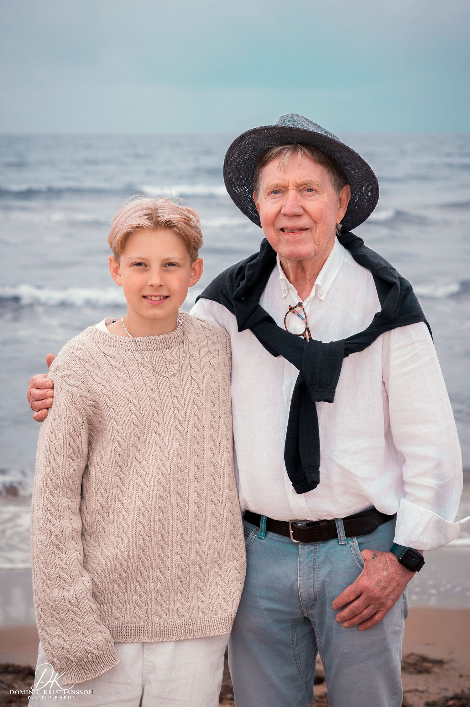 An elderly man in a hat and white shirt poses with a young boy in a cable-knit sweater on a beach.