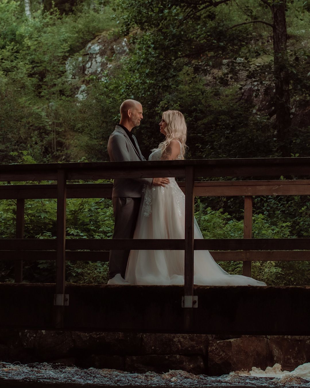 Newlyweds share a romantic moment on a wooden bridge over a stream.