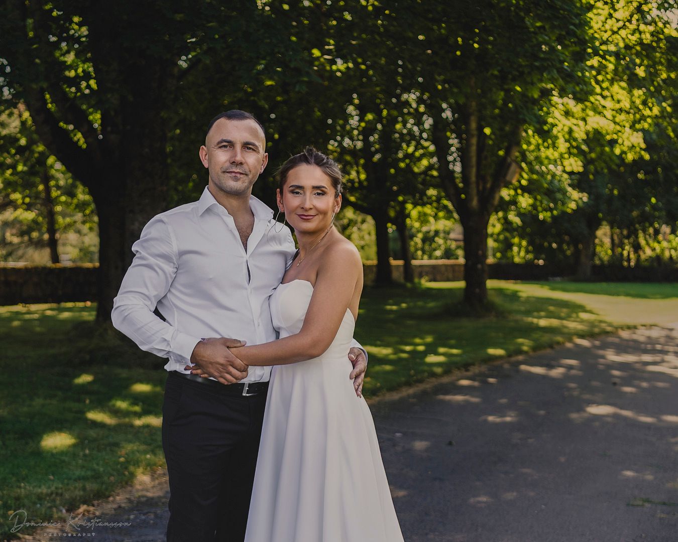 A couple in wedding attire embraces in a sun-dappled park setting during a romantic outdoor photo session.
