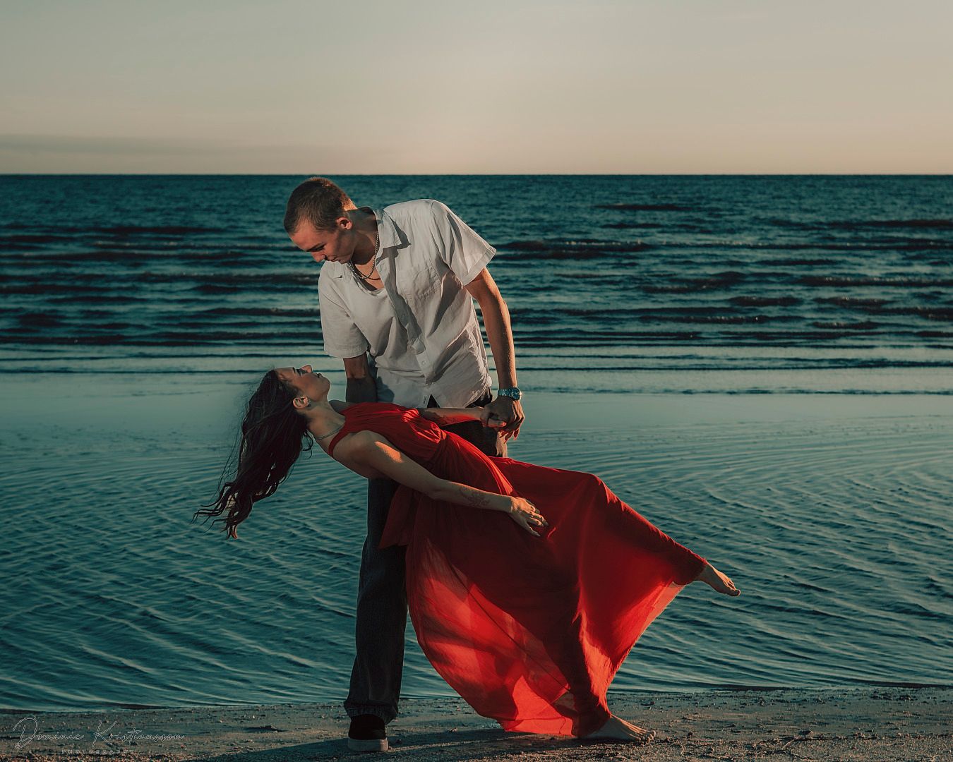 A romantic dance pose on the beach at sunset with a flowing red dress creating dramatic movement against turquoise waters.