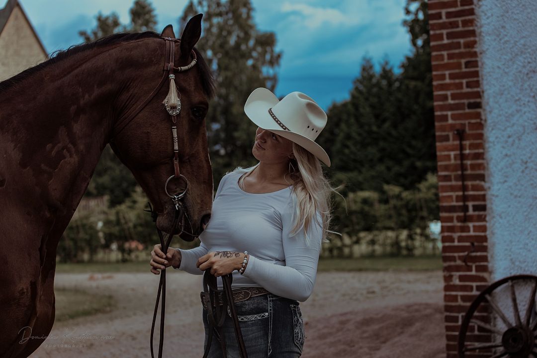 A brown horse stands beside someone wearing a cowboy hat and white top at a rustic farm setting.
