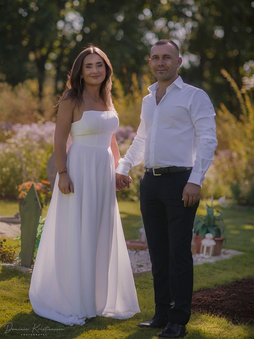 A couple poses outdoors in a garden setting wearing white and black formal attire during a photo session at sunset.