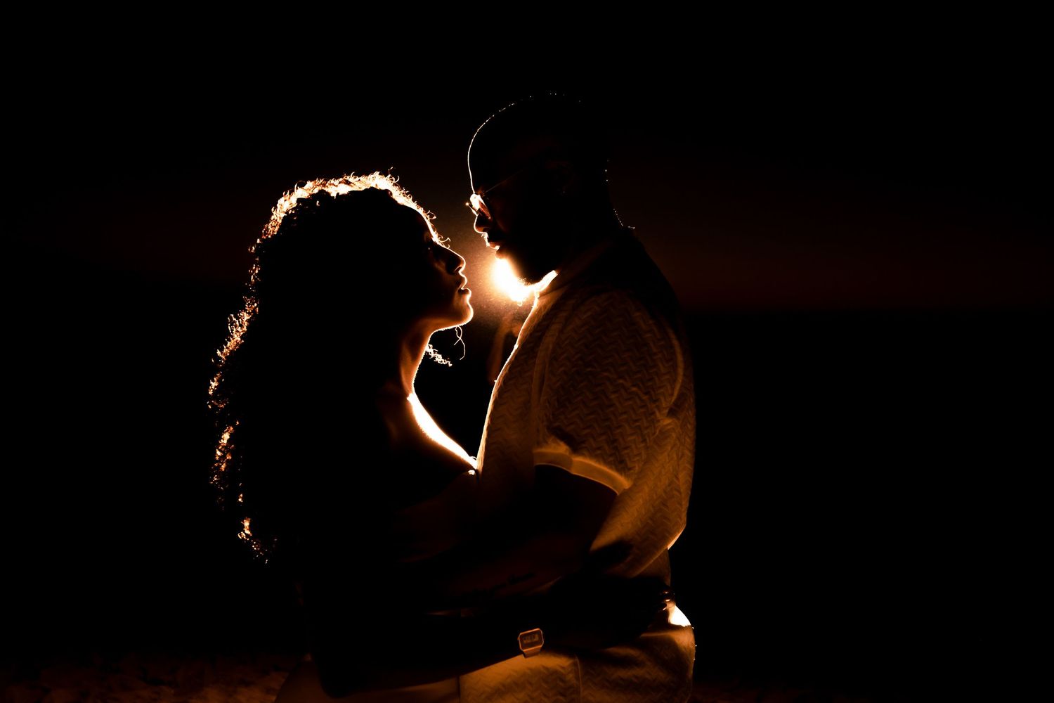 Intimate backlit portrait of couple against dark background with warm rim lighting.