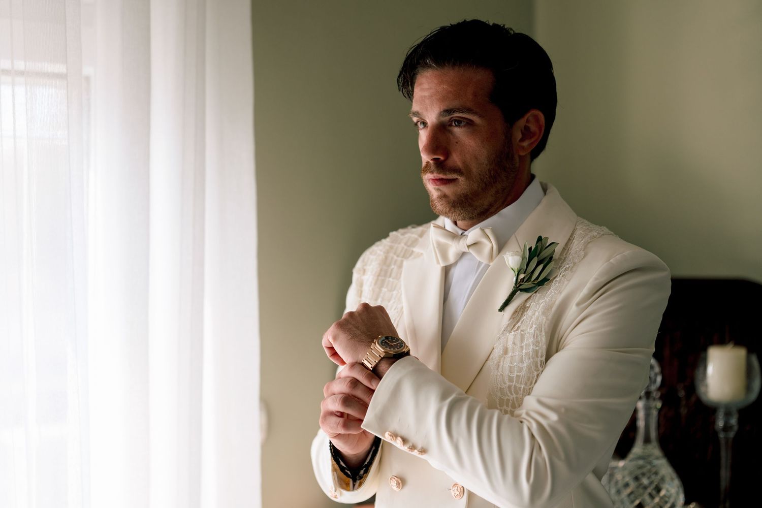 Groom in crisp white tuxedo adjusts his collar near a window.